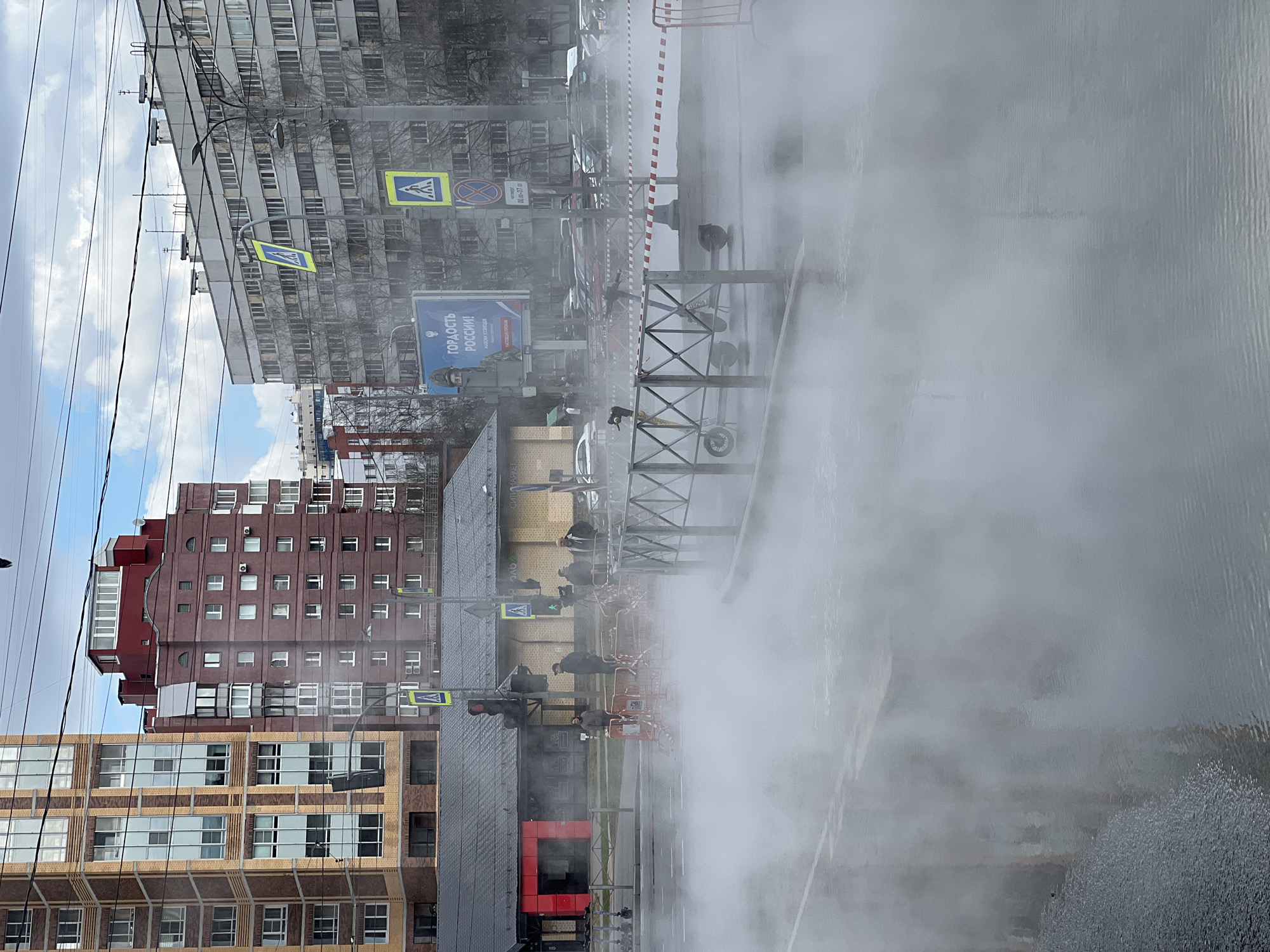 The image shows a street scene in Saint Petersburg, Russia, partially obscured by steam or vapor. Visible are several multi-story buildings with various architectural styles, including modern brick structures and older, Soviet-era apartment blocks. Street signs and traffic lights are also visible. A group of pedestrians is gathered near the steam or vapor source, which appears to originate from a utility maintenance site marked off with barricades. The street is wet, suggesting recent rain or the ongoing operation of the steam release.