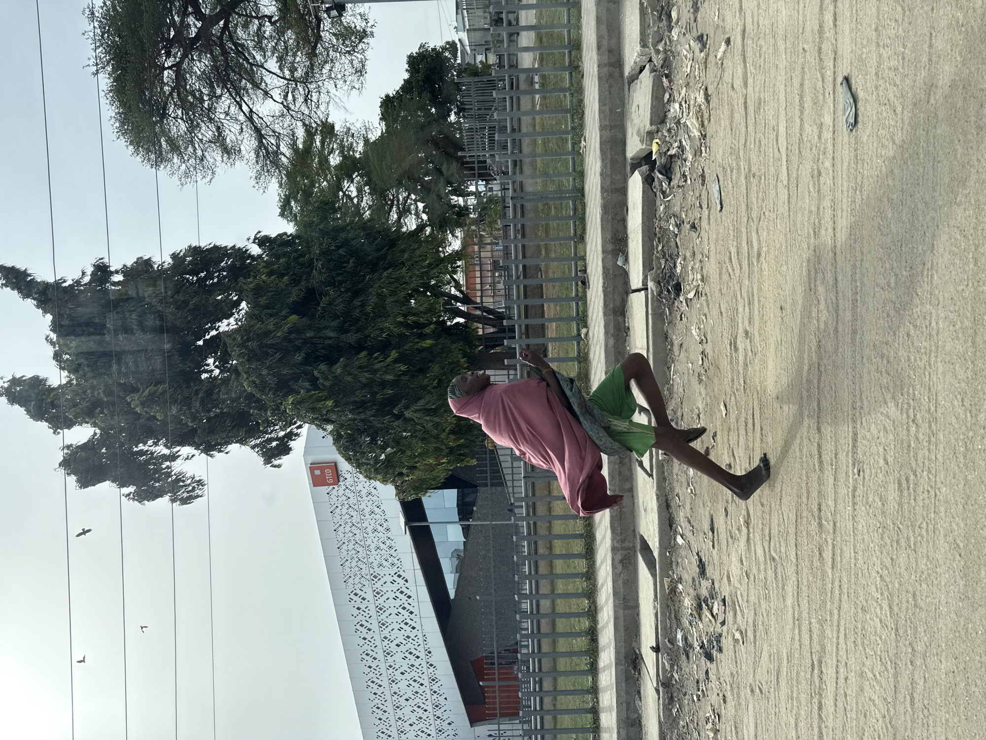 The image depicts an outdoor scene in Lekki, Nigeria, under a bright, possibly overcast or hazy, daytime sky. In the foreground, an individual is walking barefoot along a sandy or dirt path adjacent to a concrete curb. The person is dressed in a pink, flowing garment that covers their upper body and legs, with green shorts visible underneath, and a matching pink head covering.
To the left of the person, a modern white building stands prominently. Its facade is adorned with intricate geometric patterns, and a red sign on its upper right side clearly displays the text "GTCO". Surrounding the building and extending into the background are several large, lush green trees, some with dense foliage. A metal fence with vertical bars separates the sandy path from a grassy area.
Thin power lines or communication cables traverse the sky horizontally, with several small birds visible flying between them. The ground is a mix of packed sand and concrete, with some litter scattered along the edge of the curb. The overall scene captures a moment of daily life in a bustling environment.