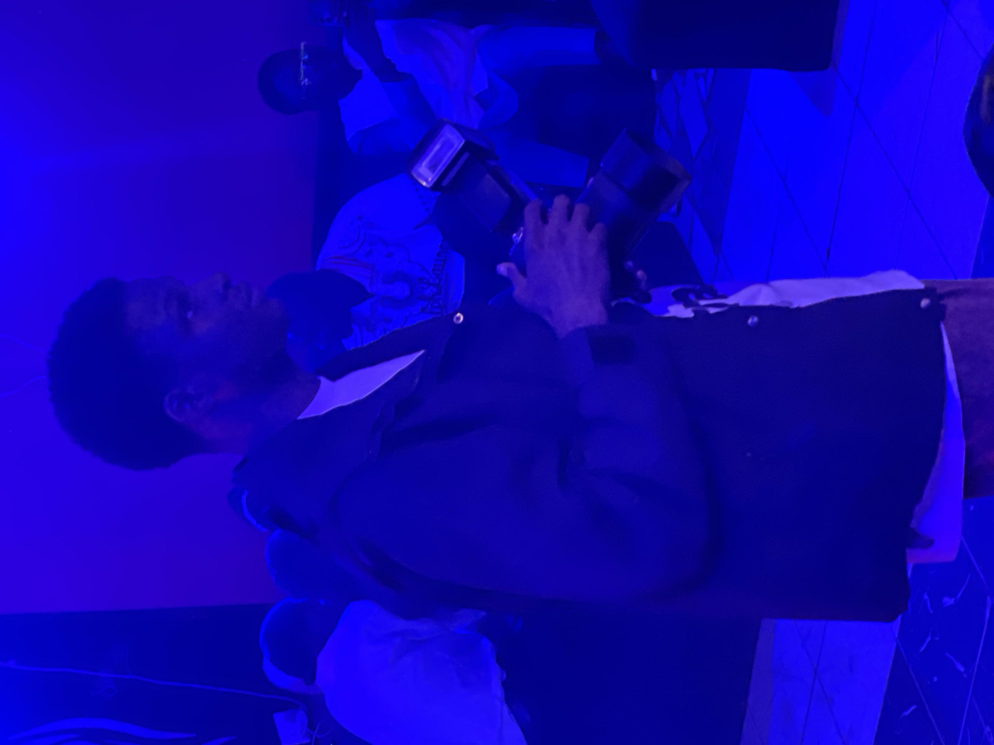 In Durumi, Nigeria, a person is holding a camera with a flash attached. The person is wearing a dark jacket over a white shirt. The environment is lit with blue lighting, casting a blue hue over the scene. Other individuals are visible in the background, also illuminated by the blue light. The flooring appears to be tiled.