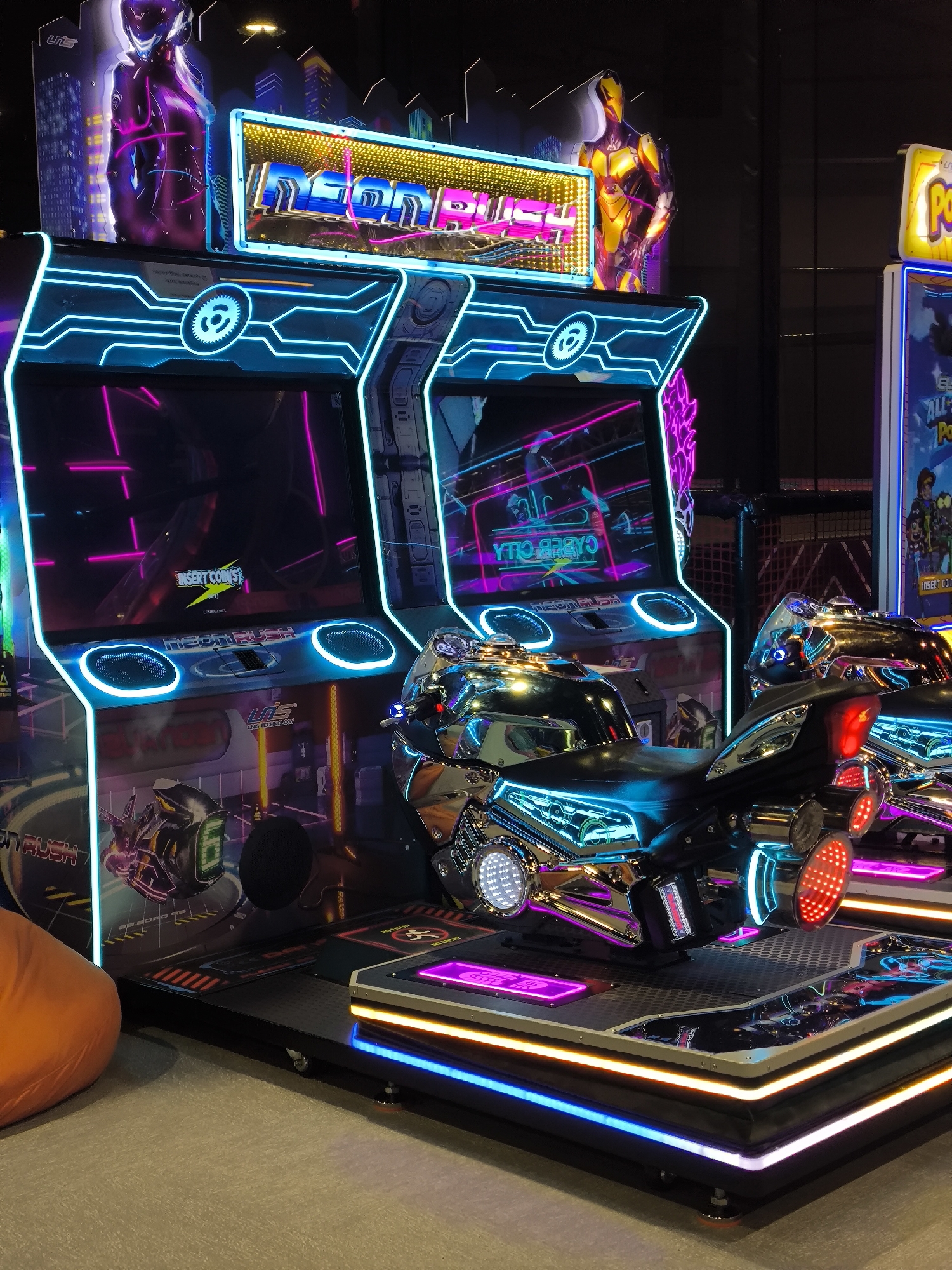 The image shows a dimly lit indoor arcade with neon-lit game machines and motorcycles. Two arcade cabinets, branded "NEON RUSH", are prominent, featuring glowing blue and pink outlines and screens displaying virtual environments. In front of the cabinets are two futuristic motorcycle game simulators with chrome finishes and extensive neon lighting in blue, red, and pink. The floor is a dark, textured surface with additional neon lighting strips in blue and yellow framing the game platforms. To the left, an orange beanbag chair is visible. In the background, other arcade machines and decor, including a large neon sign and illuminated figures, are present. The overall atmosphere is vibrant and technologically advanced. The location is identified as the city of Орловка, Russia.