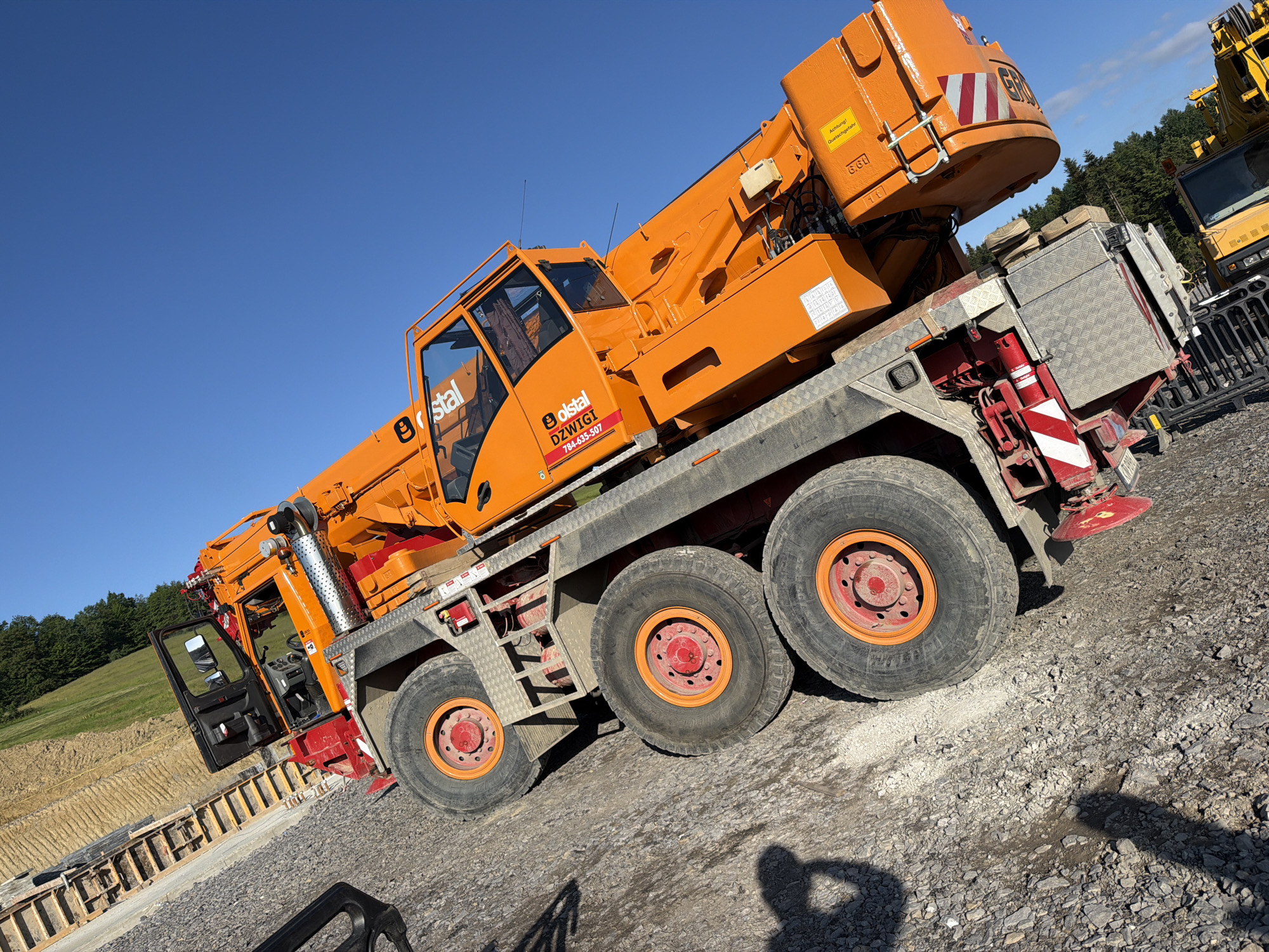 An orange and gray crane truck is parked on a gravel surface under a clear blue sky. The truck has multiple large tires and an extended boom. The crane cab is visible, with the door ajar. A company logo that reads "olstal" is visible on the side of the truck. In the background, there is a forested hillside and a construction site with wooden forms. To the right, another yellow crane truck is partially visible, as well as a black fence. The scene appears to be a construction or industrial area.