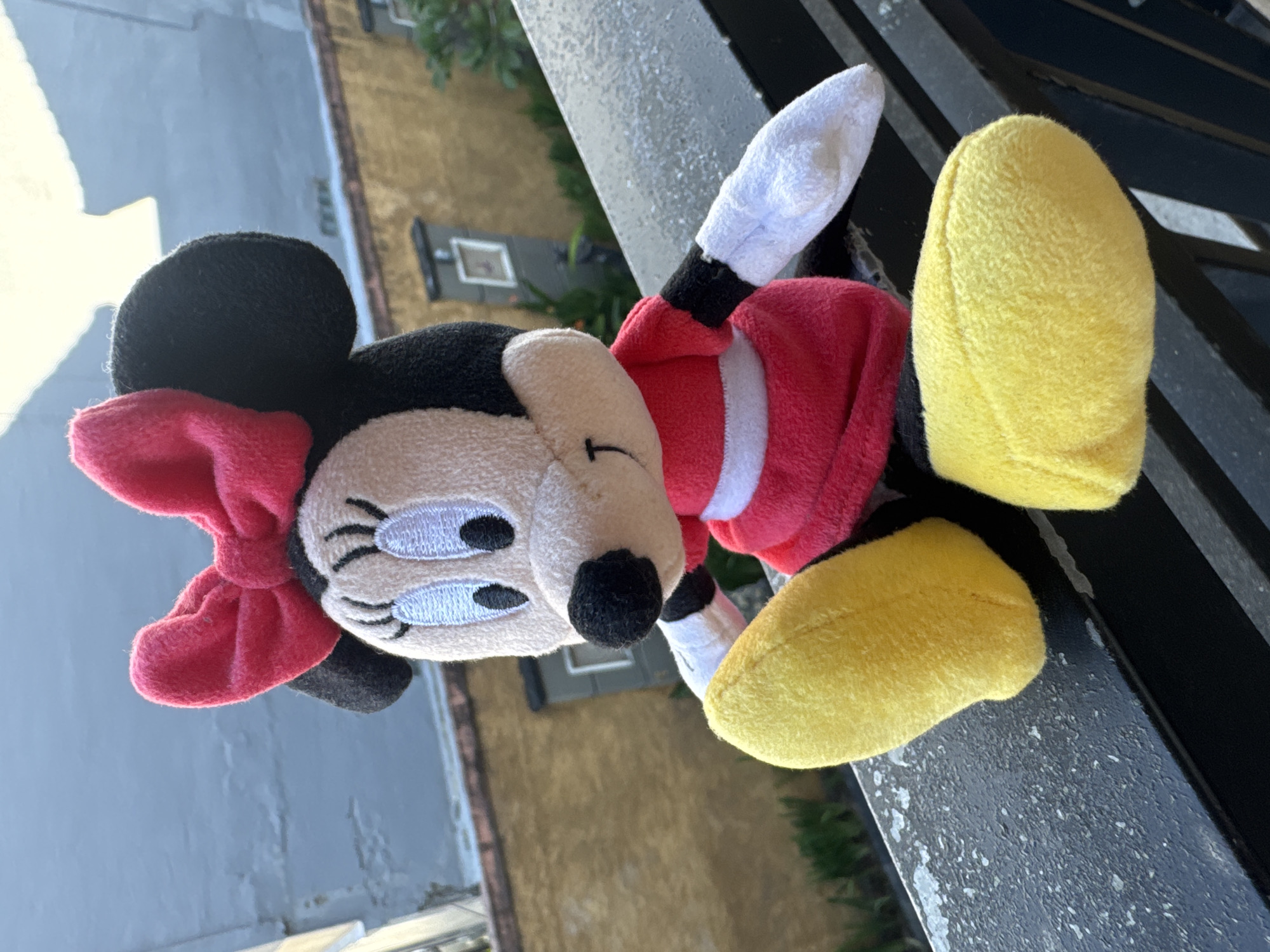 A plush toy resembling Minnie Mouse is positioned outdoors, resting on a dark-colored railing. The toy is predominantly black and white with a red and yellow dress. It has a large red bow on its head, a white face with black and blue embroidered eyes, and a black nose. Its arms and legs are white and yellow, respectively. The background shows a weathered building with windows, suggesting an urban or residential setting. The lighting indicates it is daytime, possibly sunny, given the bright colors and shadows. The overall scene is casual and appears to be a still-life arrangement of the toy in an outdoor environment.