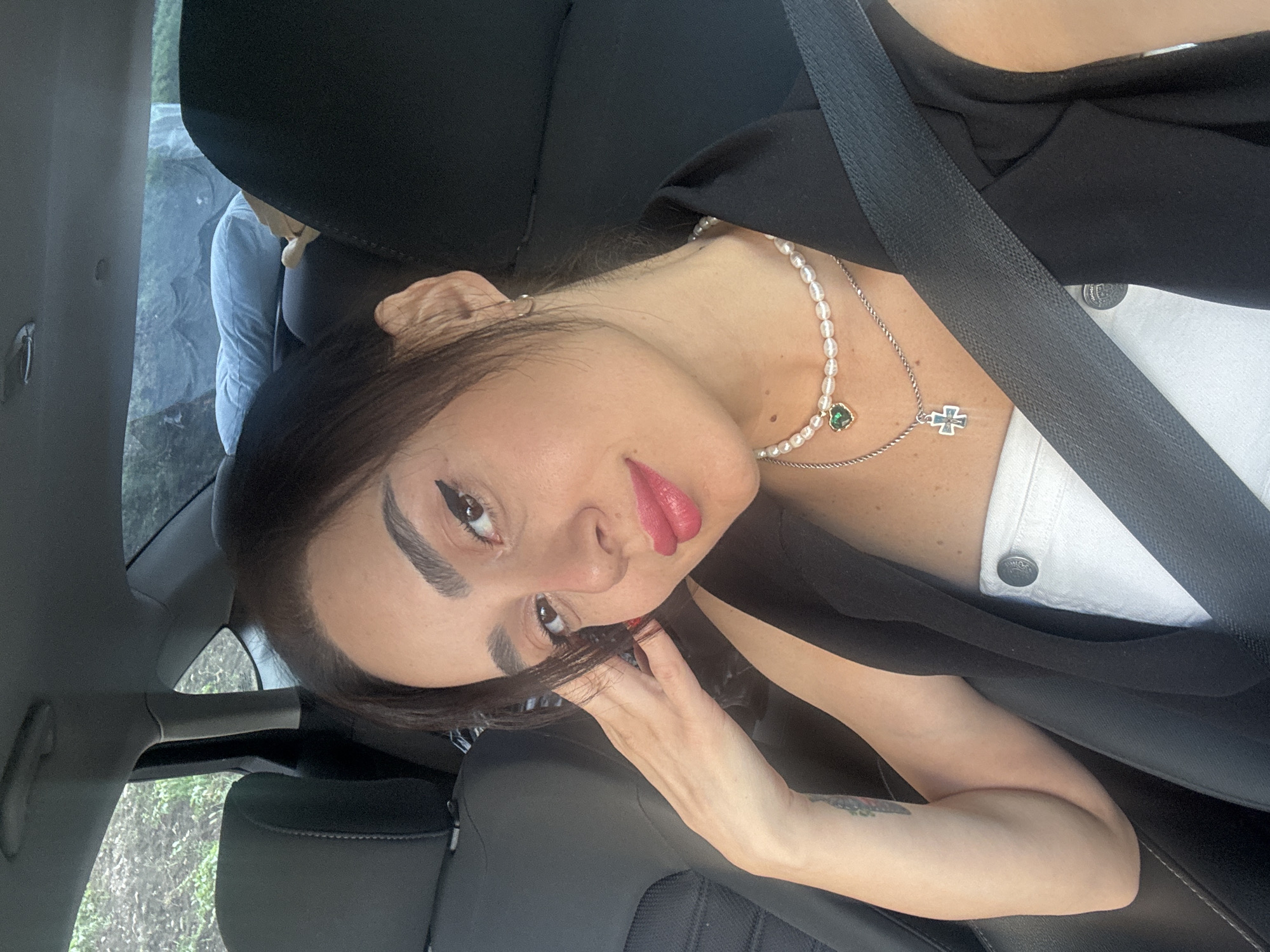 The image shows a close-up, selfie-style shot of a young woman sitting in the passenger seat of a car. She is wearing a black sleeveless jacket over a white top, and two necklaces, one with a pearl string and a green heart-shaped pendant, and another with a silver cross. She has dark hair pulled back, and is wearing noticeable makeup, including bold eyeliner and bright pink lipstick. Her left arm is resting on the back of her head, and her right hand is visible near her face. The interior of the car is visible behind her, with black seats and a seatbelt across her chest. Through the car windows, lush green foliage can be seen, suggesting the car is in a natural or semi-rural setting. The ambient light suggests it is daytime. The scene does not provide any specific visual cues to pinpoint a location within Tbilisi, Georgia.