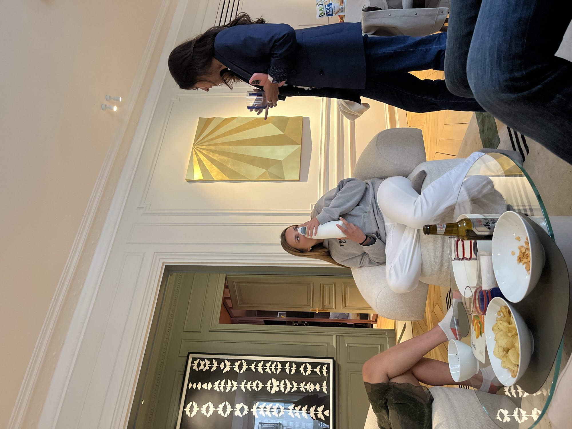 The image captures an indoor scene in Paris, France, depicting a social gathering. A woman in a blue blazer and jeans stands near a wall adorned with a gold-colored, abstract art piece. Another woman sits comfortably in a light-colored chair, holding a white bottle to her mouth. The scene includes a glass table with bowls of snacks and drinks. In the background, there is a doorway leading to another room. A dark-framed artwork hangs on the wall below the doorway.