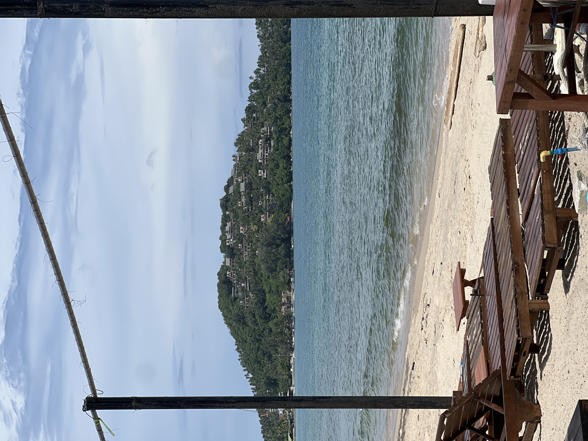 This is a medium shot, eye-level, outdoor photograph taken on a beach with a view of the ocean and a lush green hill. In the foreground, there are several empty wooden lounge chairs arranged on the sand. The sand appears light in color and wet near the water's edge. The ocean is a deep blue-green with gentle waves. In the middle ground, a tree-covered hill rises from the coastline, dotted with numerous buildings, some of which appear to be villas or resorts. The sky is partly cloudy with a mix of blue and white. Two dark, vertical poles and a horizontal pole with some wires are visible in the foreground, framing the scene.