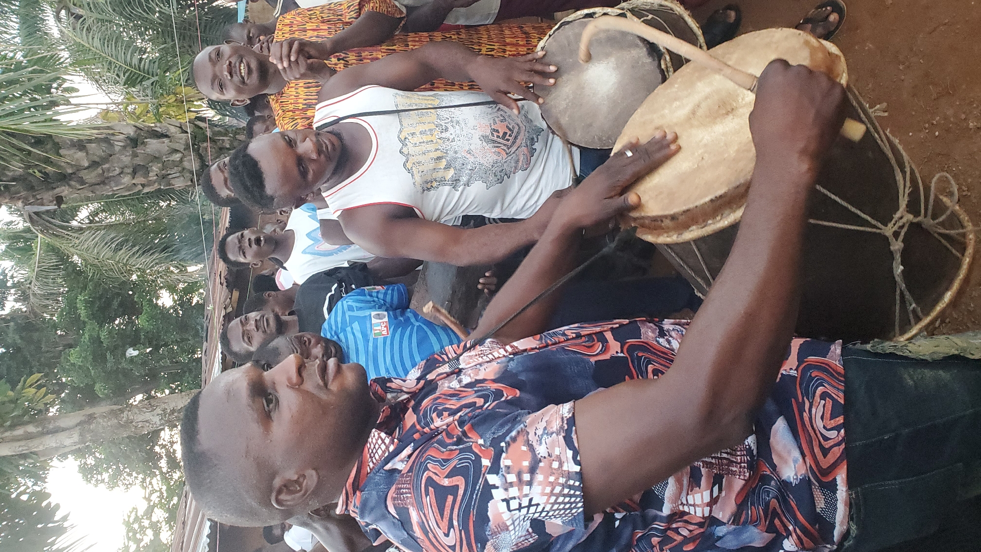 This image depicts a group of men gathered outdoors, appearing to be in Akwanga, Nigeria, engaging in a drumming activity. In the foreground, two men are actively playing drums. The man on the left is wearing a colorful, patterned shirt and dark pants, and he is positioned to the left of the frame, with his hands near a drum. To his right, another man in a white tank top with a graphic print is also playing a drum, holding a stick in his right hand.
Behind these two men, a crowd of other individuals is visible, some watching the drummers and others looking in various directions. The environment appears to be somewhat informal, with trees and foliage in the background, suggesting an outdoor setting. The ground surface seems to be dirt. The overall impression is of a cultural or celebratory gathering where music and drumming are central to the event.