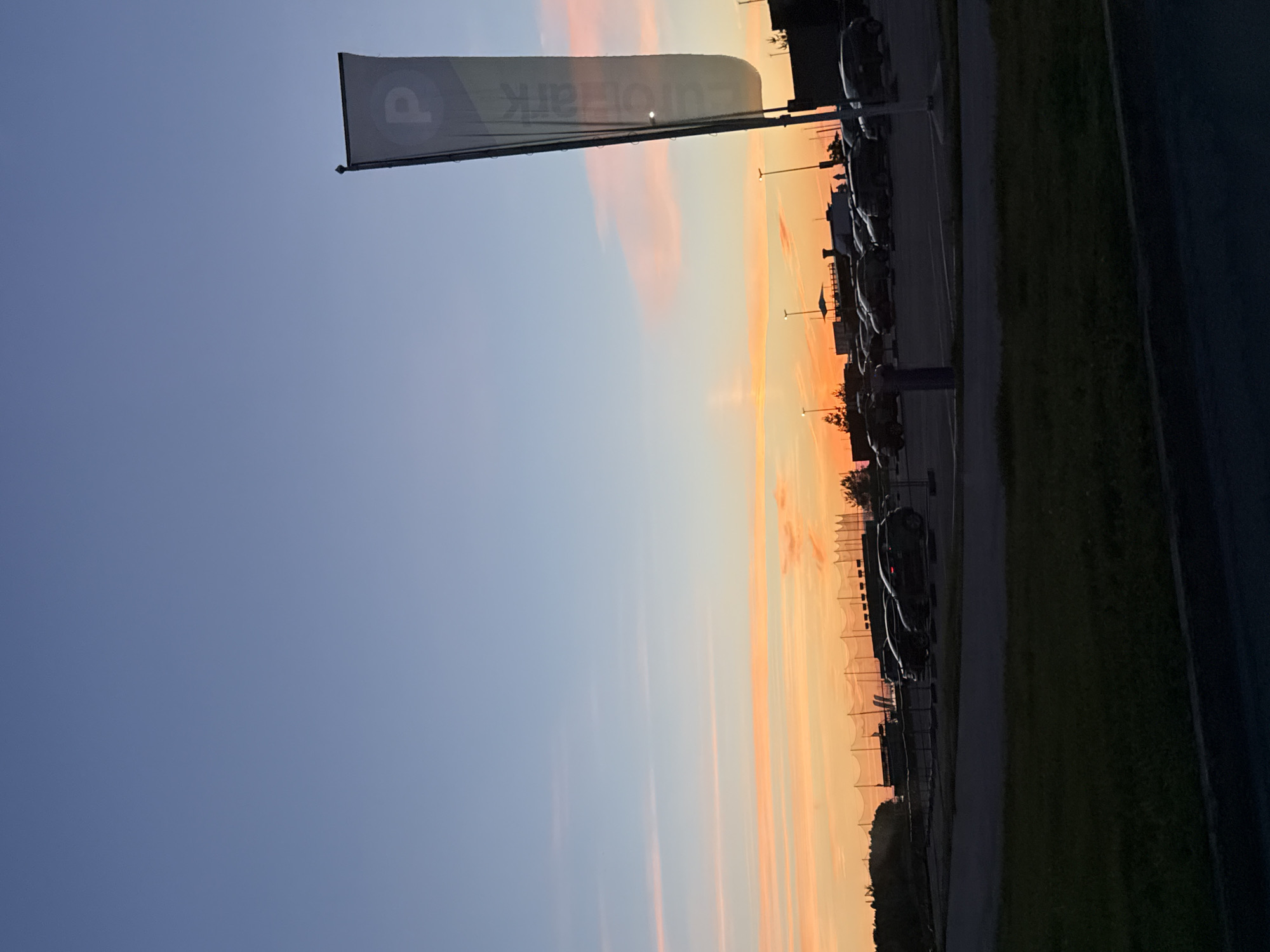 This is a wide-angle, eye-level, vertical shot of a parking lot in Tallinn, Estonia, at dusk. The sky is a gradient of blue, transitioning to orange and pink hues near the horizon, indicating sunset. A tall, dark gray flag with a white "P" logo and the word "EuroPark" printed vertically is prominently displayed on the right side of the frame. The parking lot, populated with several parked cars, is in the mid-ground, silhouetted against the colorful sky. A dark green grassy area is in the foreground, with a dark asphalt surface separating it from the parking lot. In the background, there are trees and structures, including what appears to be a driving range with a net, also silhouetted. The overall mood is serene and captures the transition from day to night.