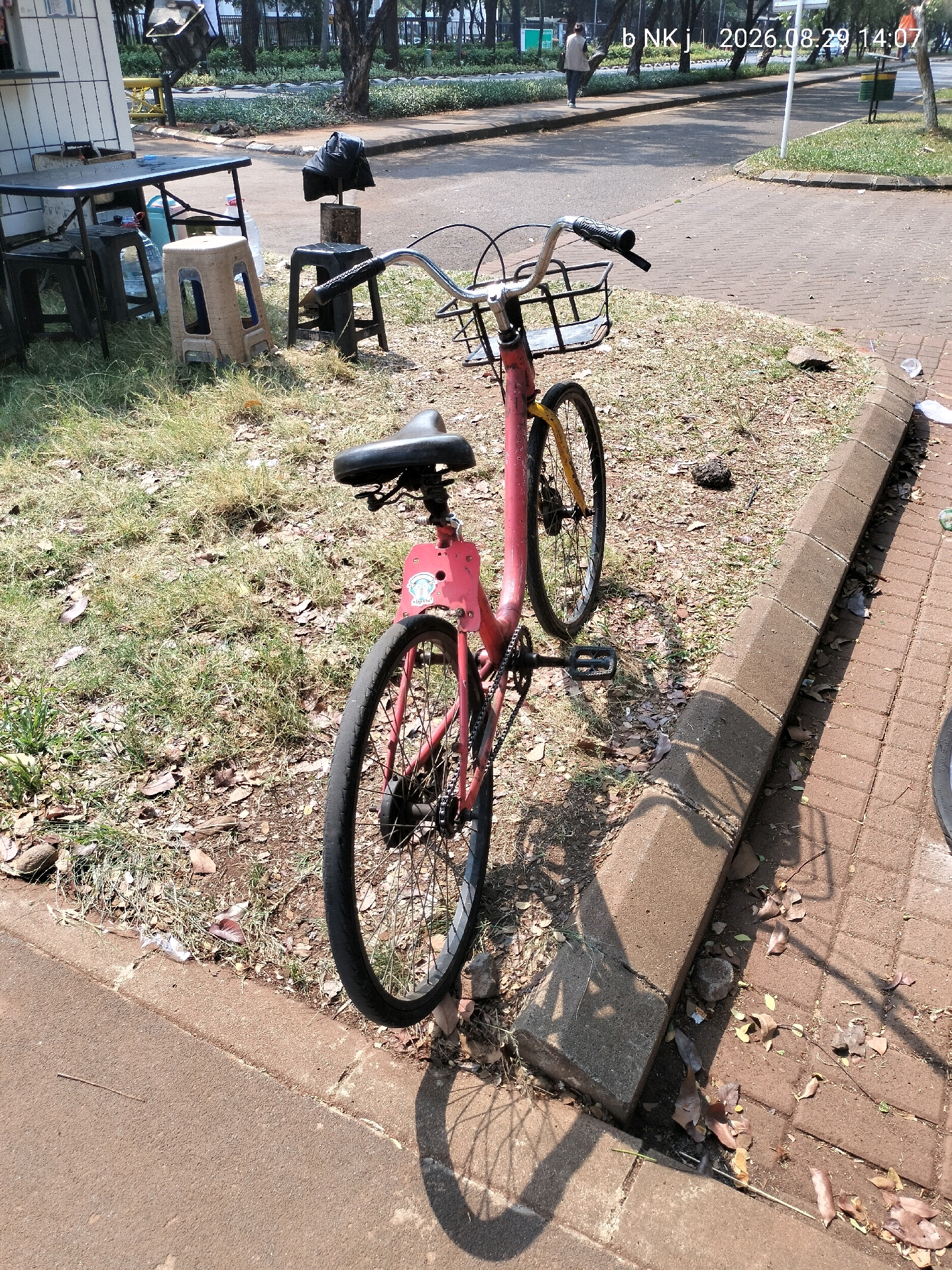 In the city of Central Jakarta, Indonesia, a pink bicycle with a black basket and seat is parked on a grassy area next to a paved walkway. The bicycle's front wheel is turned slightly to the left. In the background, there is a table and chairs set up outdoors. The ground is covered with dry leaves and grass. The sky is blue and the sun is shining. A timestamp on the image reads "2026.08.29 14:07".