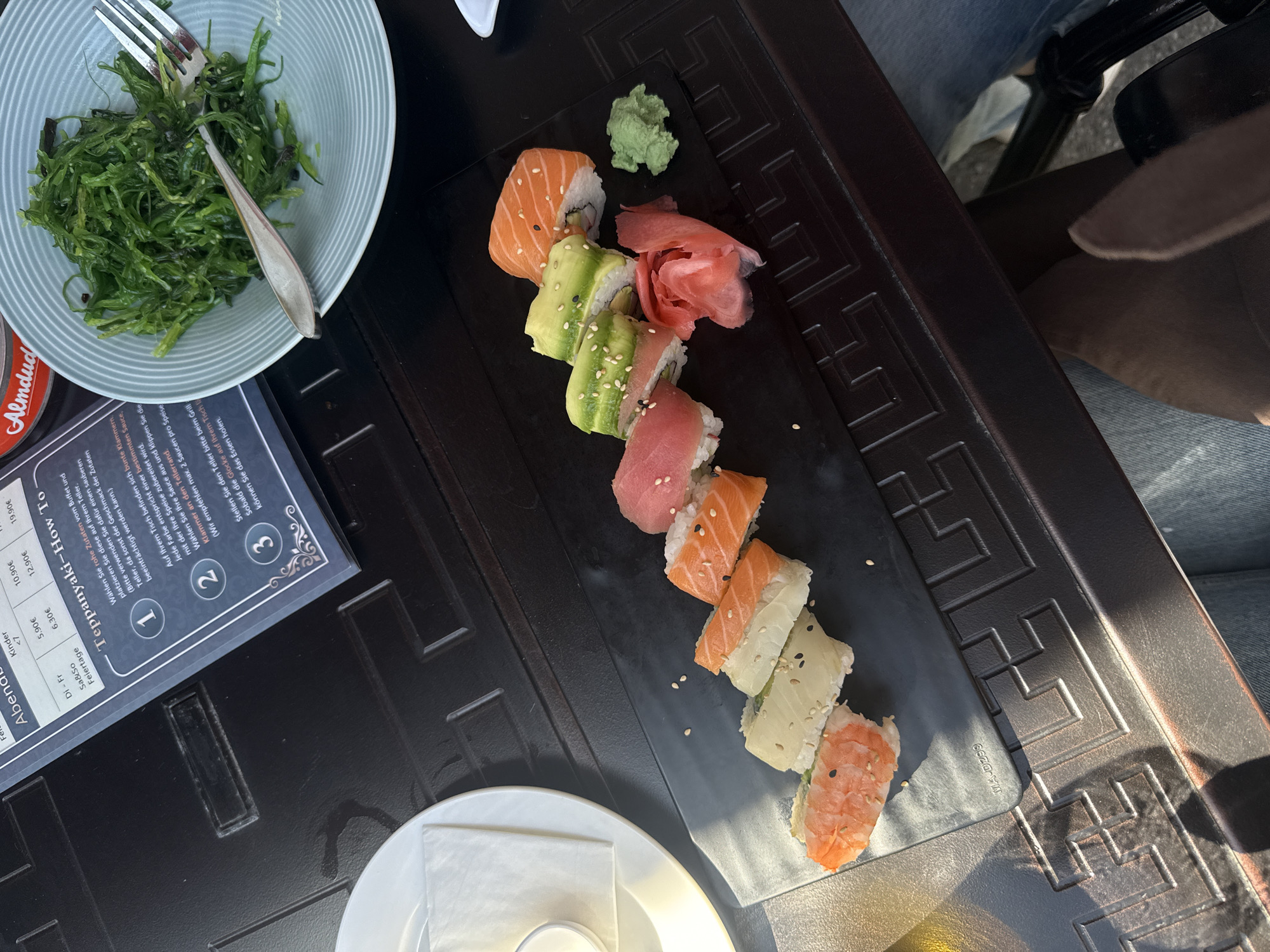 The image shows a partially outdoor dining setting, possibly in Hainburg an der Donau, Austria, based on the text visible in the picture, which features German words. The primary focus is on a sushi platter arranged on a black decorative rectangular plate. The platter includes a variety of sushi rolls with fillings like salmon, tuna, and avocado, garnished with sesame seeds. Accompanying the sushi are portions of wasabi and pickled ginger.
To the left of the sushi platter, there's a light-colored plate filled with seaweed salad, with a silver fork resting on top. Adjacent to this is a menu card written in German. In the lower-left corner, a small, round, white plate with a napkin. The image also contains a portion of someone wearing jeans, sitting at the table. The table has a textured, possibly tiled, surface. Part of a chair is visible near the top of the picture. Sunlight casts shadows and highlights on the scene.