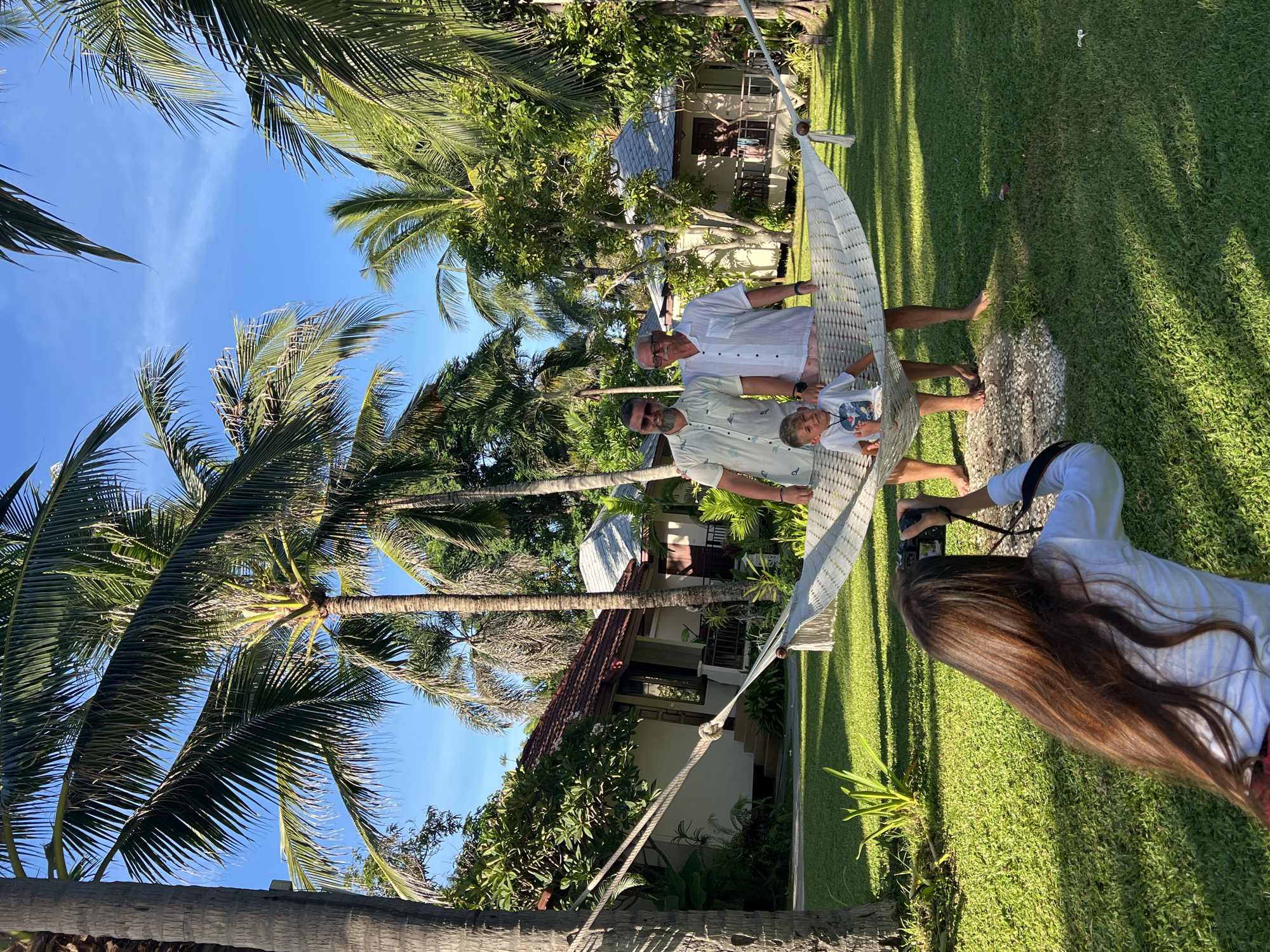 In Baan Chaweng Noi, Thailand, on a sunny day, three males are posed in a hammock in a grassy courtyard. A young boy sits in the center of the hammock, while two adult males stand on either side of him. To the left, a man with a beard and sunglasses wears a light-colored, short-sleeved patterned shirt. To his right, an older man with glasses wears a white short-sleeved button-down shirt and light pink shorts. The hammock is suspended between two palm trees. In the foreground, a person with long brown hair is bent over, holding a camera and taking a photograph of the group. The background features several bungalows with red-tiled roofs and lush green foliage, characteristic of a tropical resort. The sky is clear blue with a few wispy clouds.