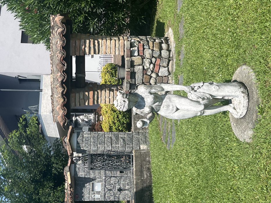 The scene depicts a residential garden in Mendrisio, Switzerland, under clear daytime conditions. A weathered, light-colored stone statue of a nude female figure, possibly depicting Eve, stands centrally in the foreground on a circular base amidst a green lawn. The statue holds an apple in its raised left hand and fabric draped over a tree stump-like element in its right hand. A white disposable face mask is positioned over the statue's eyes. To the left, a dark gray stone fence section with a decorative black wrought-iron gate and a mailbox marked "58" is visible, topped with terracotta tiles. To the right, a well structure constructed from a base of irregular stones and bricks, transitioning to uniform bricks above, features a terracotta tiled roof and an internal pulley system with rope. A potted plant in a metal bucket hangs within the well's opening. Behind these foreground elements, green and yellow-green bushes are present, along with a smaller white statue on a pedestal. In the background, a modern, multi-story building with light gray facades and balconies is partially obscured by dense green foliage from trees.