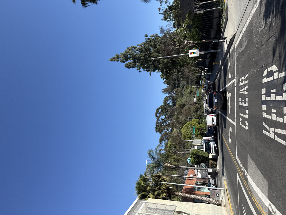 The image depicts a daytime scene at an urban intersection in Culver City, United States of America, specifically at the intersection of Jefferson Boulevard and La Cienega Boulevard. The sky is clear and blue, indicating sunny weather.
In the immediate foreground, an asphalt roadway dominates the lower half of the frame. White road markings include a "KEEP CLEAR" box, with "CLEAR" fully visible and "KEEP" partially visible. Lane division lines and arrows indicate a left-turn lane and a combined straight-ahead/right-turn lane.
Traffic occupies multiple lanes, appearing to be stopped or moving slowly. Vehicles include a dark-colored SUV in the foreground, a white delivery van, and a white United States Postal Service (USPS) truck on the left side, along with numerous other cars extending into the midground. A green traffic light is visible on the left side of the intersection, while a red traffic light is visible on the right side.
To the left of the roadway, a multi-story building with a light-colored, possibly stucco or concrete exterior featuring a grid pattern is visible. Signs near this building indicate "EMERGENCY ENTRANCE" and "PATIENT ENTRANCE" with directional arrows. Several tall palm trees are present on the left, alongside other assorted green foliage.
To the right of the roadway, a paved sidewalk runs parallel to the street. A street lamp post is prominent, attached to which is a traffic signal displaying a red light and a sign for a "PEDESTRIAN ENTRANCE." An electric scooter is parked upright on the sidewalk near the curb. Various trees, including a tall evergreen conifer and several palm trees, line the right side of the street.
The background features a prominent, densely vegetated hill, part of the Baldwin Hills, covered with numerous trees and shrubs under the bright blue sky. No individual people are distinctly visible, but vehicles imply occupants.
