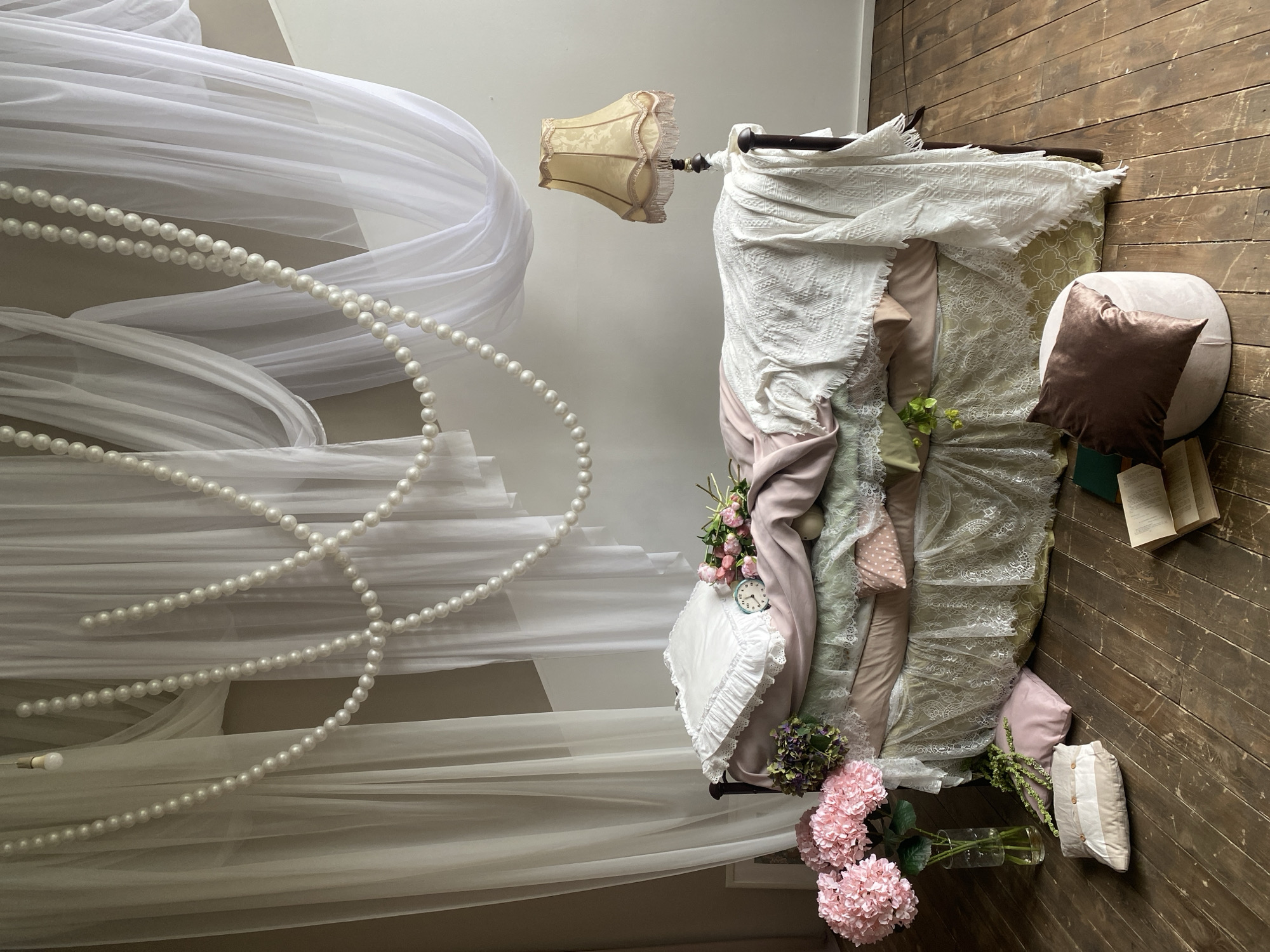The image depicts an indoor setting with a romantic and whimsical aesthetic. The scene features a bed adorned with layers of fabrics including lace and light pastel colors. The bed has a canopy draped with white, sheer fabric and strings of pearls. A vintage-style lamp with a patterned lampshade sits near the head of the bed.
To the right of the bed, on a wooden floor, is a round ottoman with a brown velvet pillow. A book is open on the floor near the ottoman. The scene is embellished with floral arrangements, including pink hydrangeas and other mixed flowers, some in vases.
There is no indication of location or whether the scene is from Yekaterinburg, Russia. The image contains no people, actions, or interactions.