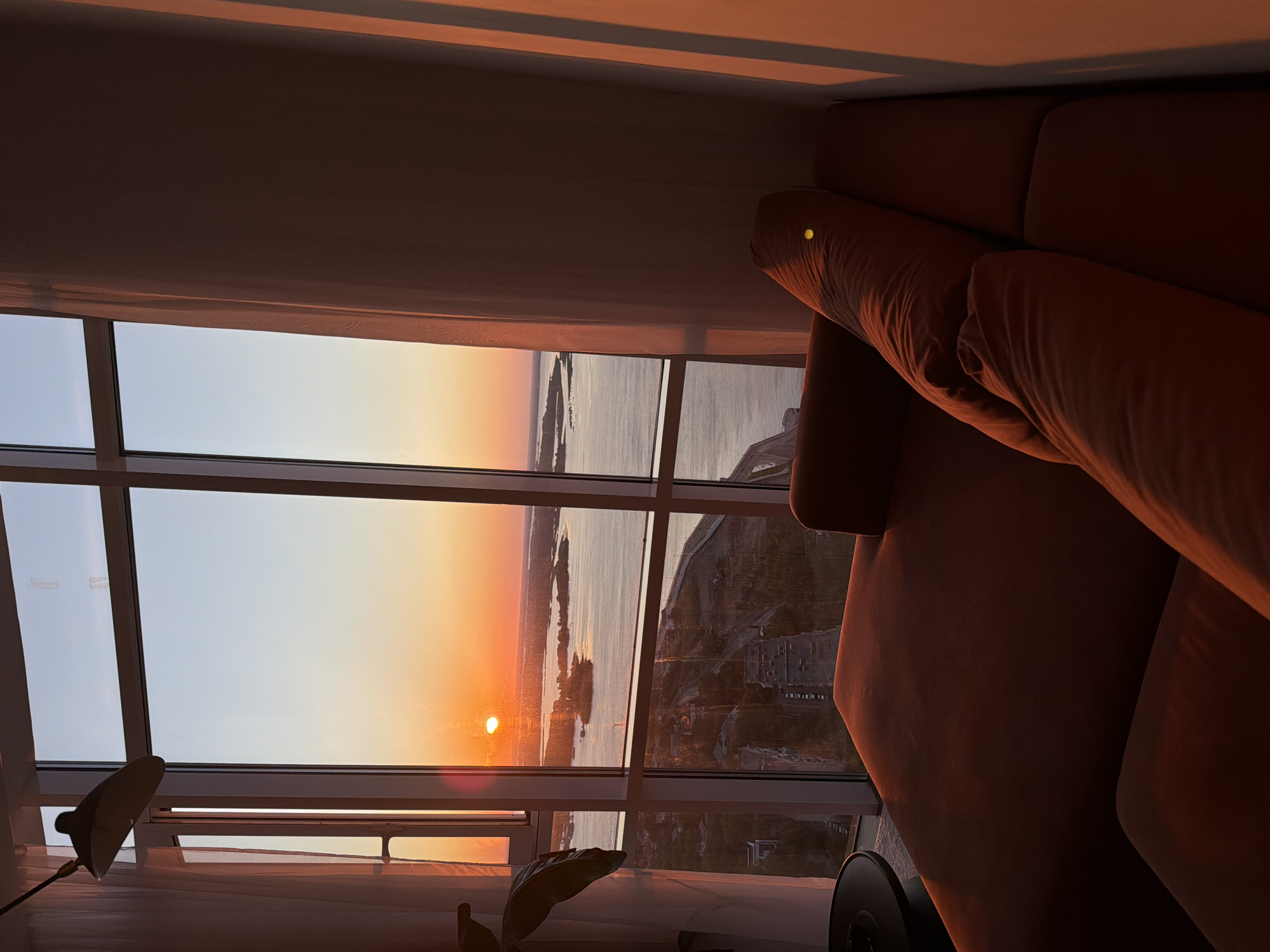 A sunset view from a high-rise apartment in Saratov, Russia, is framed by large windows. The setting sun casts a warm, orange glow across the sky and the Volga River. In the foreground, a plush, orange sectional sofa occupies the lower half of the image. The view through the windows reveals islands in the river and the cityscape of Saratov below, including buildings and roads. A ceiling-mounted lamp is visible in the upper left corner, and sheer white curtains hang on the left and right sides of the windows.