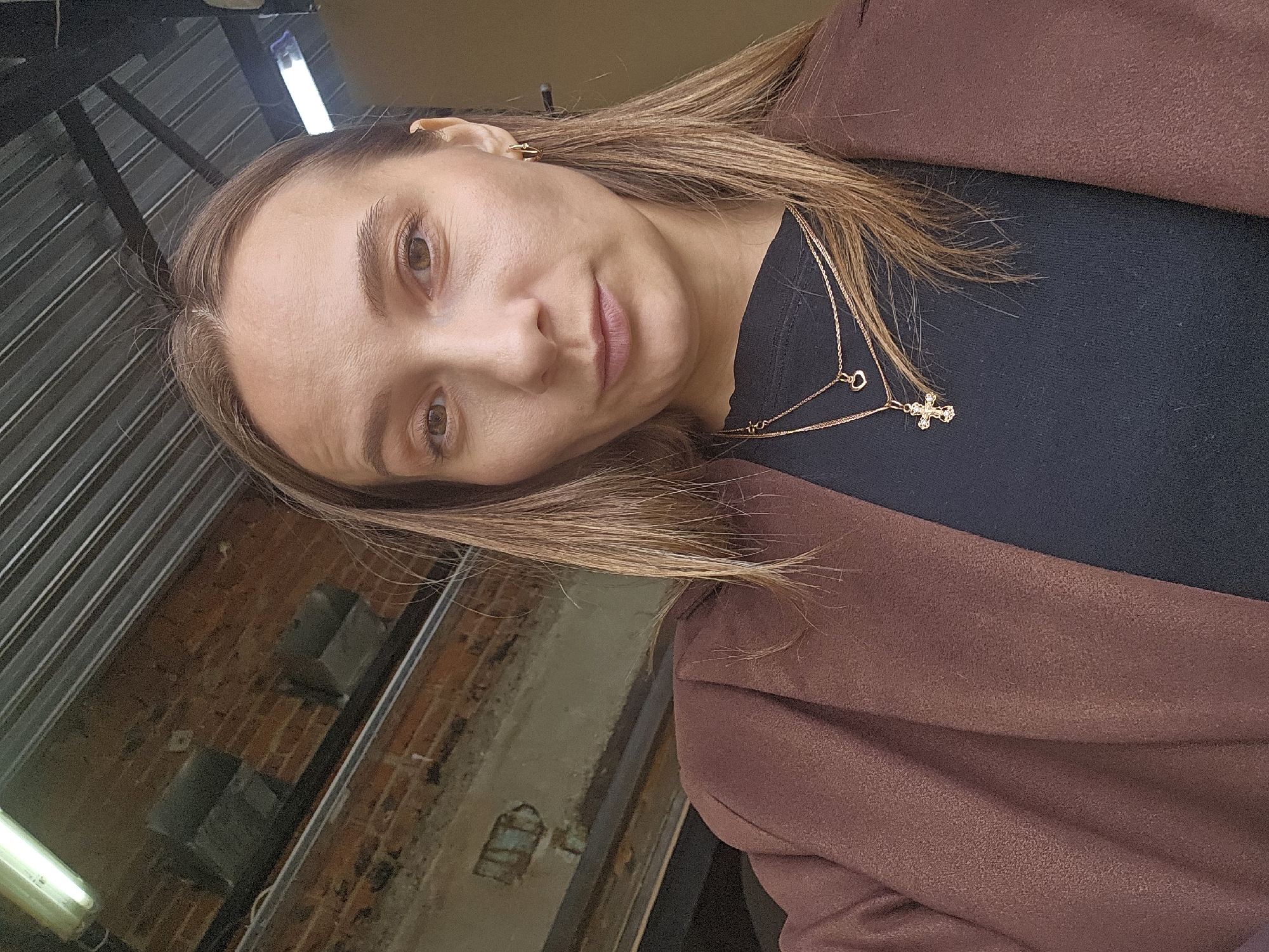 A close-up, tilted portrait shows a woman indoors, looking directly at the camera with a neutral expression. Her head is tilted to the right, and she has light brown hair that falls over her shoulder. She is wearing a dark top, possibly black, and a brown or maroon jacket or coat. Visible accessories include small gold hoop earrings and two delicate gold necklaces. One necklace features a small heart pendant, while the other displays a jeweled cross.
The background suggests an industrial or rustic interior setting. To the left, there are vertical dark metal beams or corrugated panels. Below these, and extending across the right, is an exposed brick wall, showing signs of wear and various shades of red and brown. A fluorescent light fixture is partially visible in the bottom left corner. The overall lighting appears to be ambient indoor light. There is no visible text in the image. The scene conveys a casual, candid moment within what appears to be a repurposed or uniquely designed space, consistent with urban settings like Chaikovsky, Russia.