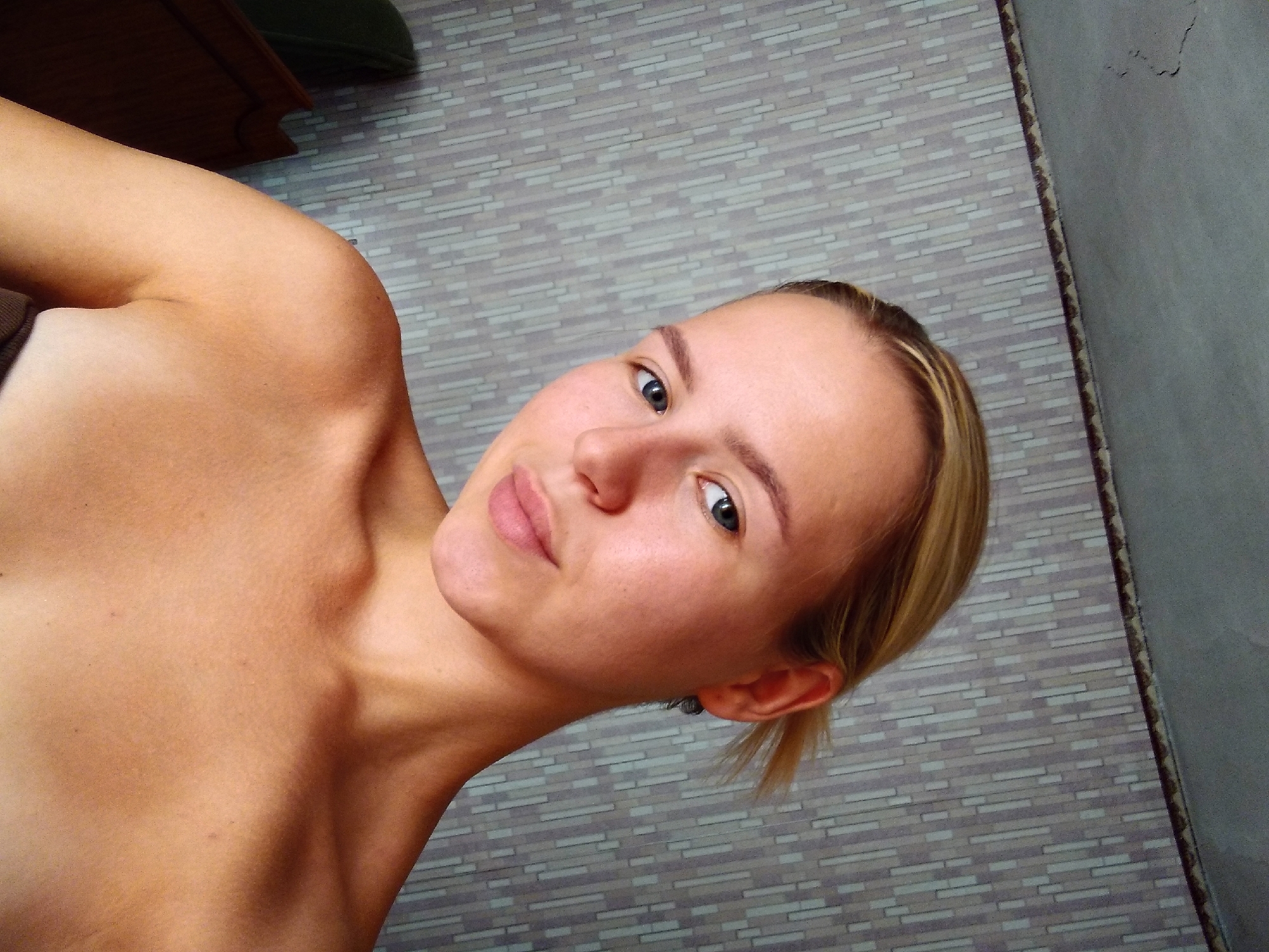 A young woman with blonde hair pulled back is shown from the shoulders up, looking directly at the camera. She has blue eyes and light skin. She is not wearing any visible clothing above the shoulders. The background consists of a patterned wallpaper. The image appears to be taken indoors, with a section of a ceiling visible at the top. There is no indication of the location in Radisne, Ukraine.