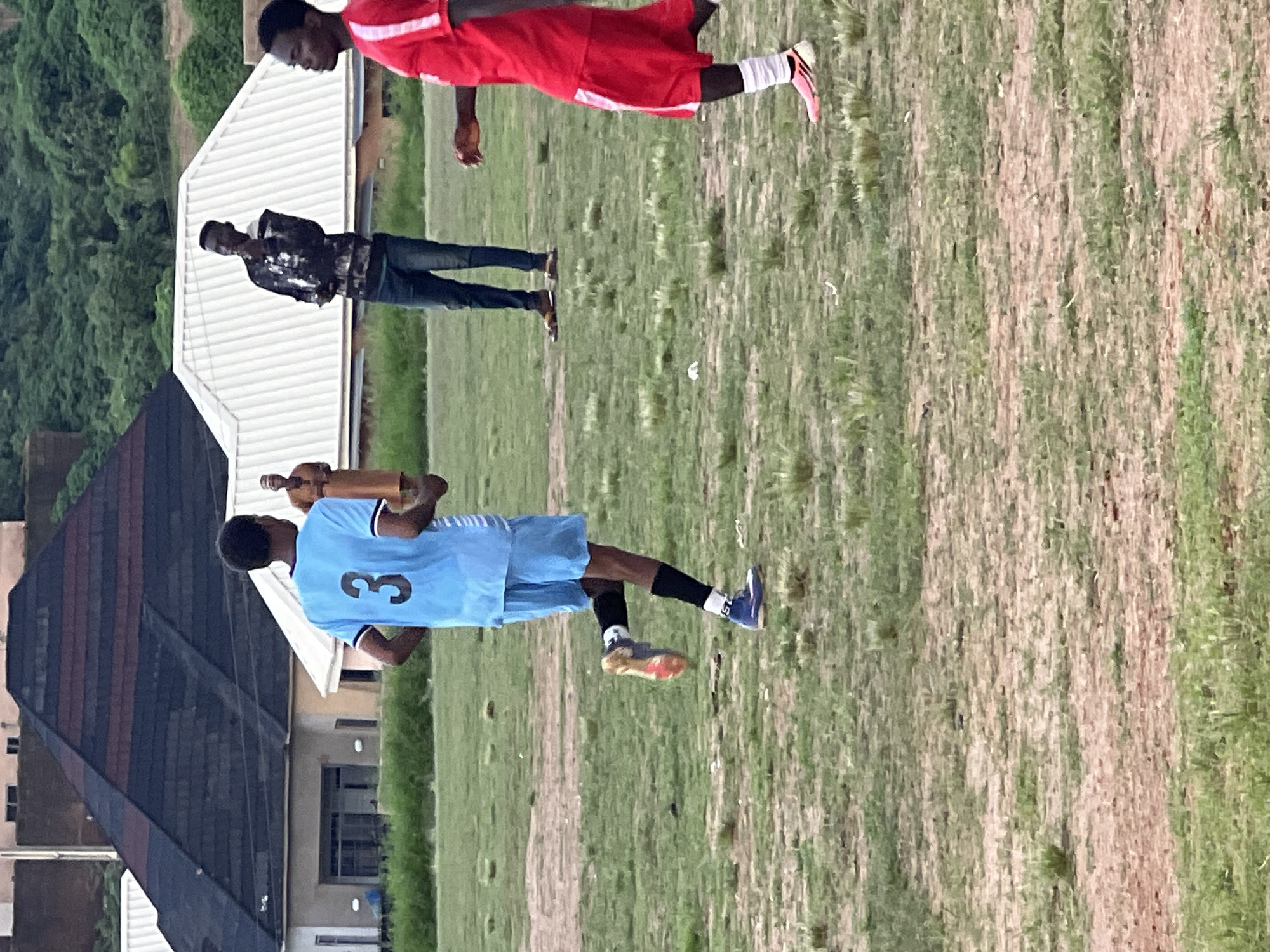 The image depicts an outdoor scene in Jos, Nigeria, possibly a sports field, with a group of young men engaged in activity. In the foreground, a person wearing a light blue jersey with the number '3' and matching shorts is captured mid-stride, suggesting they are running. They have black socks and blue athletic shoes. To the right, another individual in a red jersey and shorts is visible, also in motion, with white socks and pink athletic shoes. Further in the background, two other individuals are present. One, wearing a patterned dark shirt and blue jeans, stands with a hand near their chin. Another person, dressed in a light brown traditional garment and cap, is also standing. The environment consists of a dry, grassy field with patches of brown earth. In the background, a large, dark, pyramid-shaped roof structure dominates the scene, with white-roofed buildings behind it. Hills covered in lush green vegetation rise in the distance under an overcast sky. The overall atmosphere appears casual and communal, likely an informal sporting event or practice.