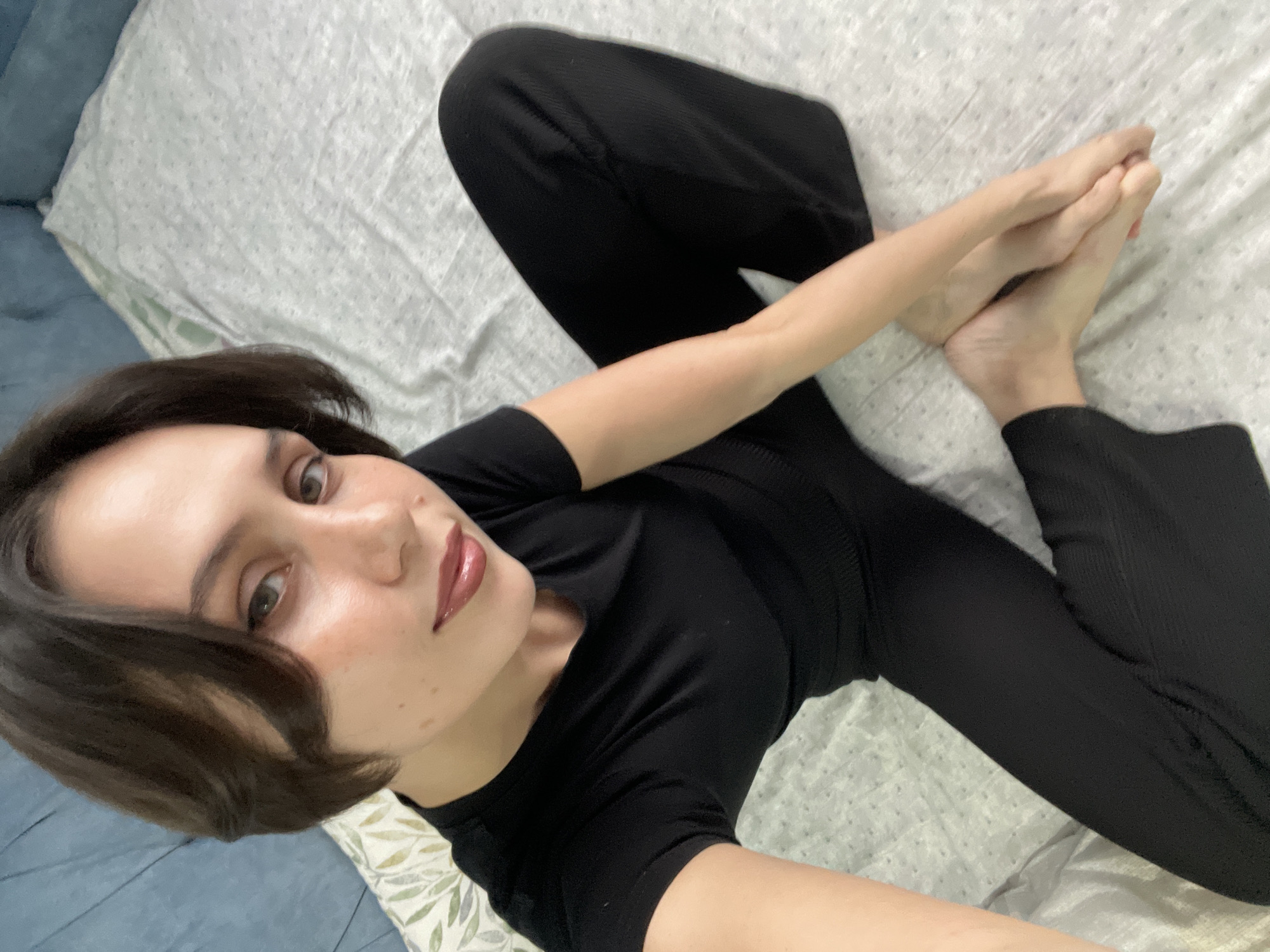 A woman with short brown hair and wearing a black shirt and pants is lying on a patterned bedsheet. Her legs are crossed, and she is holding her feet with her hands. She is looking at the camera. The scene appears to be indoors, possibly in a private residence. The photograph's location is not discernable from the image.