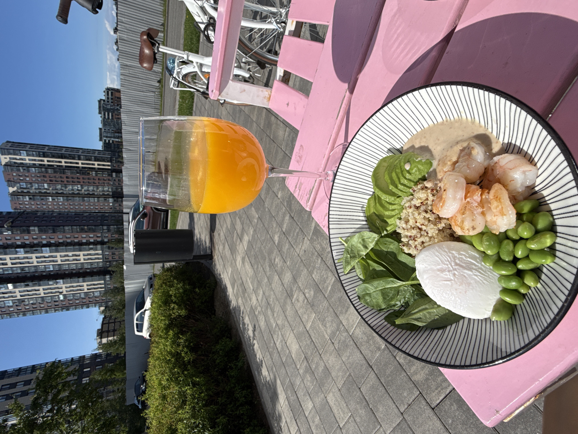 The image captures an outdoor dining scene on a bright, sunny day, likely at a cafe or restaurant patio. In the foreground, a vibrant light pink wooden table or bench serves as the surface for a meal.
On the right side of the table, a stylish bowl with thin black and white stripes holds a colorful and healthy dish, resembling a poke bowl. The ingredients are clearly visible: several cooked shrimp, a fan of neatly sliced avocado, a portion of quinoa, fresh green spinach leaves, bright green edamame beans, and a perfectly poached egg. A creamy, light-colored sauce is drizzled over some components, notably near the shrimp and quinoa.
To the left of the bowl, a stemmed wine glass holds a bright orange beverage, presumably orange juice.
The background reveals an urban environment. A white bicycle with a brown seat and handlebars is partially visible behind the table, suggesting a casual setting. The ground is paved with light grey rectangular tiles. Further back, tall, modern-looking apartment buildings with numerous windows rise against a clear blue sky, indicating a city or residential area. Green bushes line parts of the paved area and the base of some buildings. A few cars are visible parked in the distance, partially obscured by the foliage and structures.
The strong sunlight casts distinct shadows on the pink table and paved ground, suggesting it is daytime, likely morning or early afternoon. No people are visible in the image, nor is any text discernable. The scene conveys a sense of peaceful enjoyment of a meal outdoors in an urban landscape.
