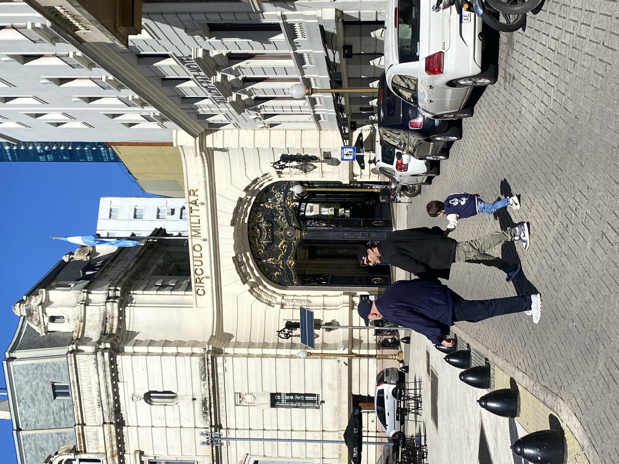 A bright daytime urban scene in Buenos Aires, Argentina, features a grand, cream-colored building on the left and a street with pedestrians and parked cars on the right. The multi-story building exhibits intricate classical architecture with decorative moldings, balconies, and a prominent, ornate dark entrance gate embellished with gold. The words "CIRCULO MILITAR" are displayed in capital letters on its facade. An Argentine flag, with its light blue and white stripes, is visible flying from a pole on the building's roof.
On the right, a paved street is lined with several parked vehicles, including a white Volkswagen Gol with a partially visible license plate ("AE 69...") and a motorcycle. Black bollards line the sidewalk, where three people are present. In the foreground, two adult men are walking away from the camera; one wears a dark blue jacket and cap, the other a black jacket and cap. Further ahead, a small child in a blue jacket with a white emblem also walks away.
The setting is sunny under a clear blue sky, indicated by strong shadows, suggesting midday or early afternoon. Street furniture includes a streetlight with a solar panel, and a blue parking sign with a wheelchair symbol. Additional visible text includes an inverted "HOTEL" sign on a building across the street and "TRUFFA" on a black cover near the bollards.