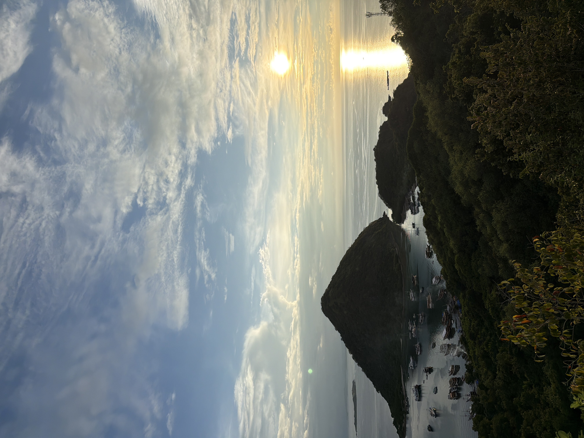 The image shows a waterscape near Labuan Bajo, Indonesia, with a predominantly clear sky filled with wispy clouds. The sun is visible over the water, creating a glare. Dark, green, tree-covered islands rise out of the water, with boats anchored in the coves. A few structures, possibly power lines or towers, are visible on the horizon.
