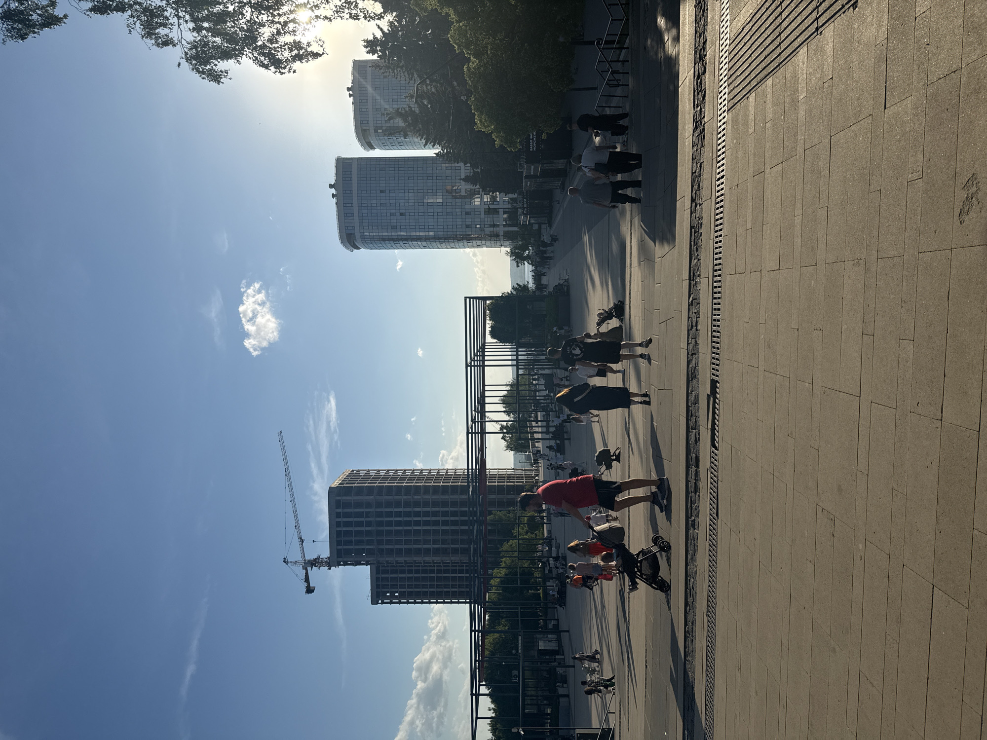 The image depicts a public space in Izhevsk, Russia. The scene features a wide, paved pedestrian walkway with several people. Some are walking individually, while others are pushing strollers. In the background, there are several buildings, including a high-rise structure under construction with a crane visible, and a couple of apartment buildings. The sky is light blue with some scattered clouds. The image is taken during the daytime, and the overall lighting suggests it's either morning or late afternoon. A set of metal structures are placed along the walkway, possibly for artistic purposes or providing shade. There are trees and other greenery between the walkway and the buildings. The area seems to be a popular place for people to stroll and enjoy the outdoors.