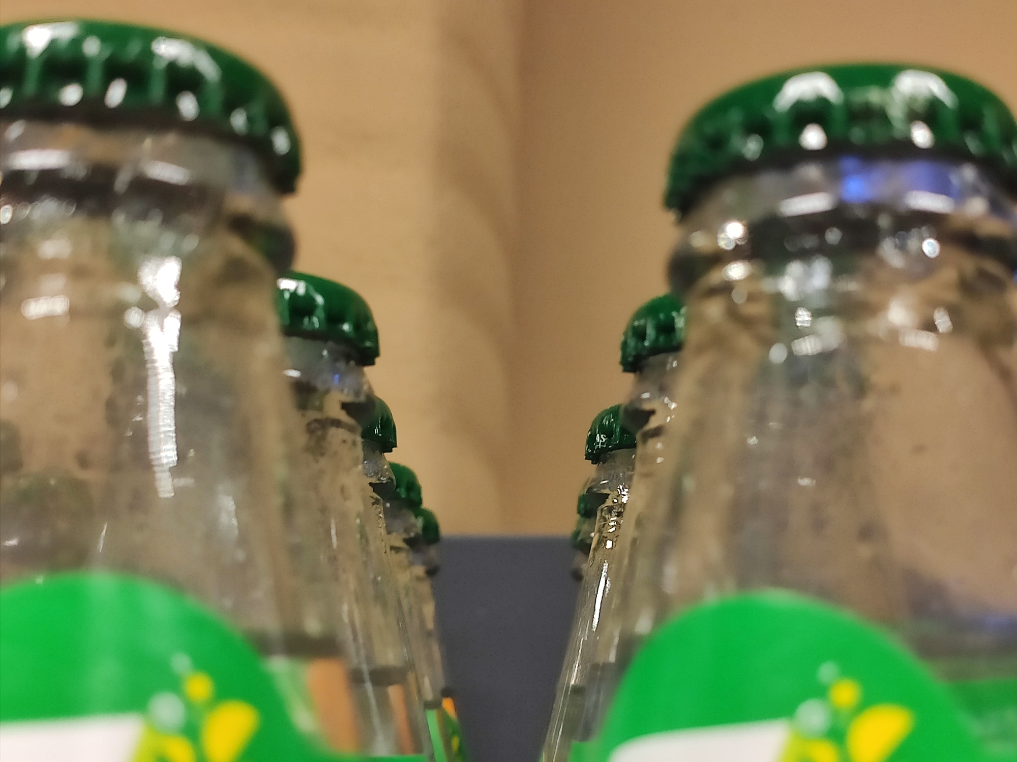 A close-up, low-angle shot focuses on the green bottle caps of several glass soda bottles. The bottles are arranged in two parallel rows, creating a narrow channel between them. The glass of the bottles is clear and appears to have some condensation or water droplets on its surface. Some of the bottles have prominent green labels at the bottom of the frame, which are partially blurred due to the shallow depth of field. The background is a blurred, light brown, suggesting a shelf or wall. The overall lighting is bright, with reflections visible on the glass surfaces, creating a glossy effect. The image does not contain any people, animals, or specific landmarks that would help determine a location within Tashkent, Uzbekistan.