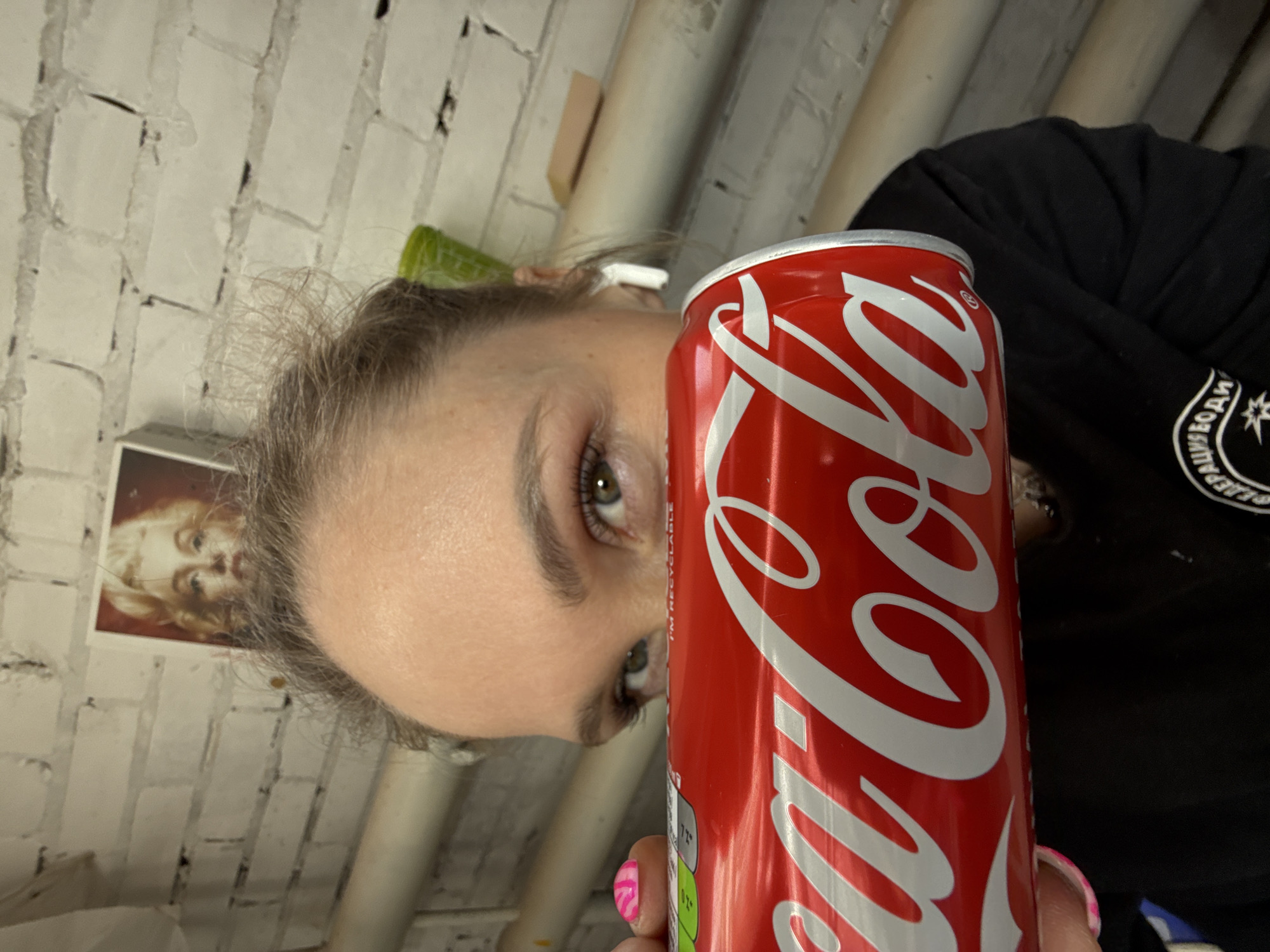 A person, identified as female, is positioned in the foreground, holding a red Coca-Cola can horizontally across their lower face, obscuring the mouth and nose. The individual's light-colored eyes are directed forward, and their forehead shows light brown hair. A white wireless earbud is visible in the right ear. The left thumb, with pink and white patterned nail polish, is seen gripping the can. The Coca-Cola can features the prominent white "Coca-Cola" logo on a red background, with "I'M RECYCLABLE" and "200ML" text visible on the upper left. The individual wears a black long-sleeved garment with a partial white logo displaying Cyrillic text "ФЕДЕРАЦИЯ ВОДИ" (Federation of Water) on the lower right. The background consists of a white-painted brick wall exhibiting texture and wear. A light-colored horizontal pipe runs across the mid-ground. A framed image of Marilyn Monroe is mounted on the wall above the person's head. A green object and a rectangular light brown object are also visible on the wall and pipe in the upper right. The scene is located in Chaikovsky, Russia.