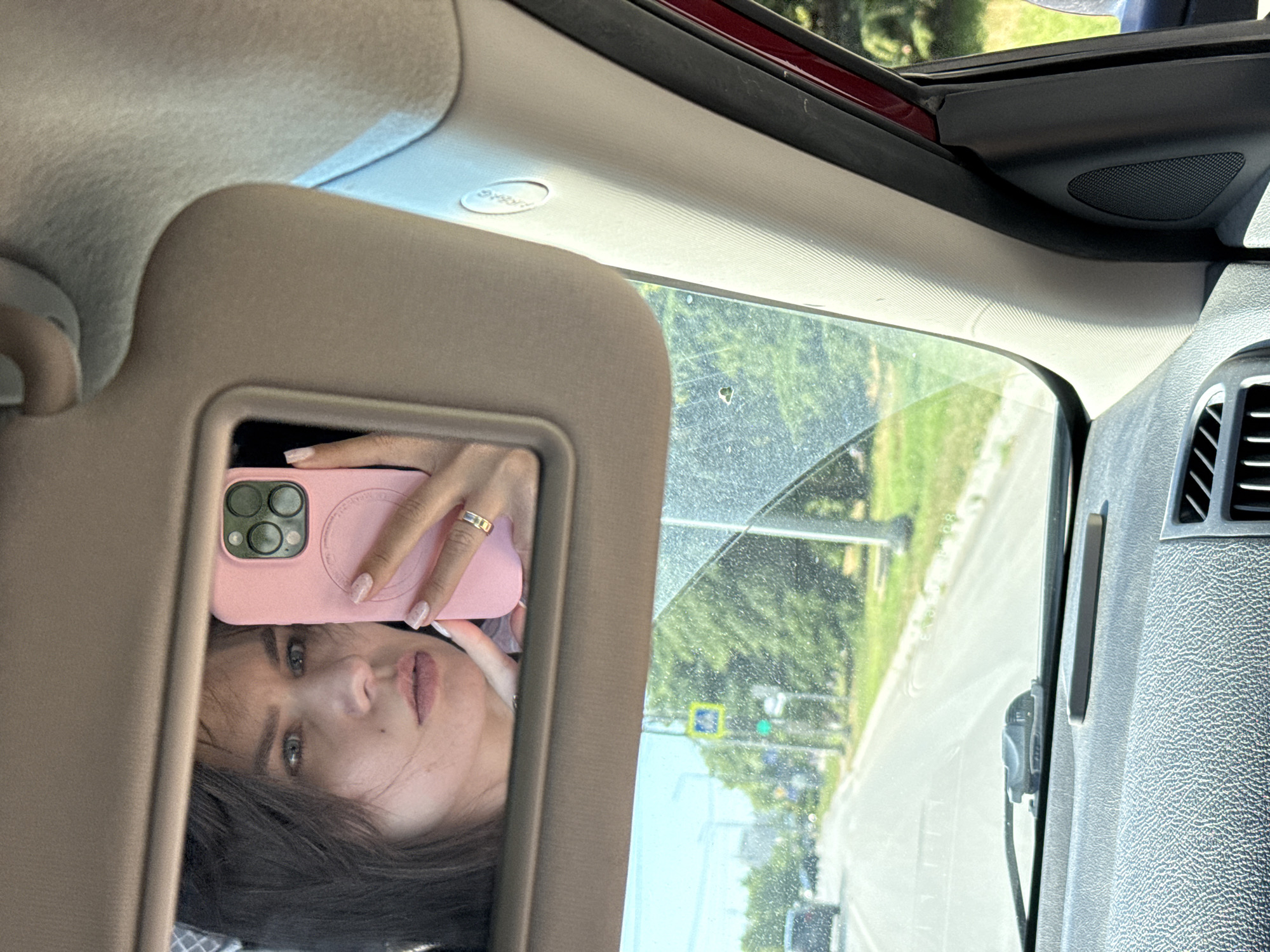 This media file shows a selfie taken inside a vehicle. The primary subject is a young woman reflected in the vanity mirror of the car's sun visor. She is holding up a pink smartphone, presumably to take the picture. She has long, dark hair that partially obscures her face, and her eyes are looking directly at the camera. She is wearing a ring on her finger. The background reflected in the mirror shows a street scene with trees and a road, suggesting it is daytime and likely a sunny day, given the brightness. The interior of the car is visible, including the sun visor and part of the dashboard. There is a visible "AIRBAG" label on the car's interior ceiling. The overall mood appears casual, as if she is documenting her journey or a moment during a car ride.