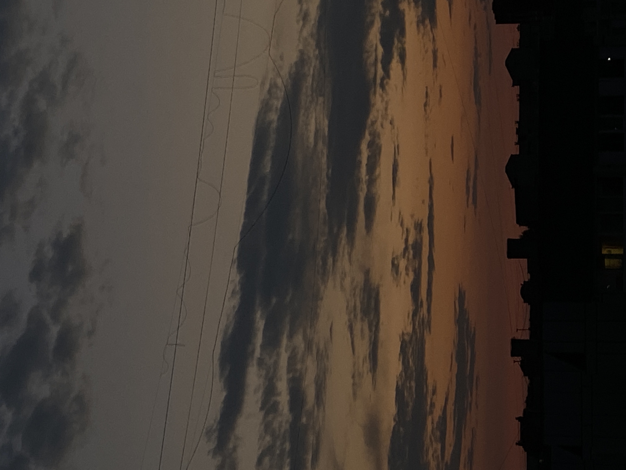 A silhouetted cityscape is visible against a dusky sky. The sky displays a gradient of colors, transitioning from a deep orange near the horizon to a muted gray at the top, with layers of dark clouds scattered throughout. Several thin wires, possibly power lines, stretch horizontally across the upper portion of the frame. The buildings in the foreground are indistinct silhouettes, their shapes suggesting a mix of structures with various rooflines and some appearing to have box-like protrusions. A single illuminated window is visible near the bottom right of the image. The scene appears to be captured during twilight or early dawn.