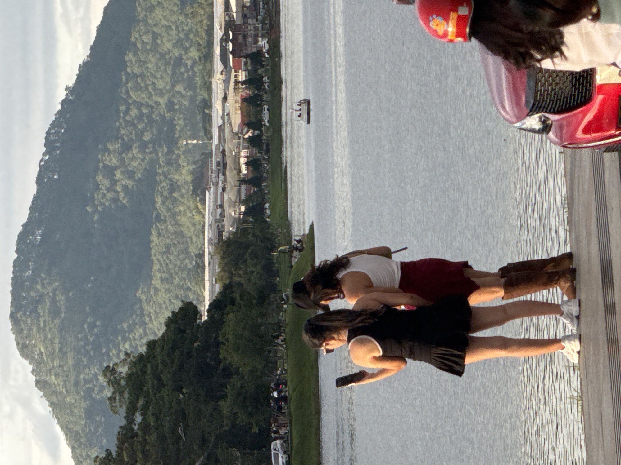 In Da Lat, Vietnam, two women stand by Xuan Huong Lake, their backs to the camera. The woman on the left, wearing a black tank top and a black pleated skirt, holds a smartphone in her left hand. She is also wearing white sneakers. The woman on the right wears a white tank top and a maroon skirt, paired with brown knee-high boots. A red vehicle, possibly a car, is partially visible in the foreground on the right. In the background, across the lake, a town with buildings and greenhouses is nestled at the base of a lush, tree-covered mountain. A small boat with people on board can be seen in the middle of the lake. The sky is overcast, suggesting a diffused light.