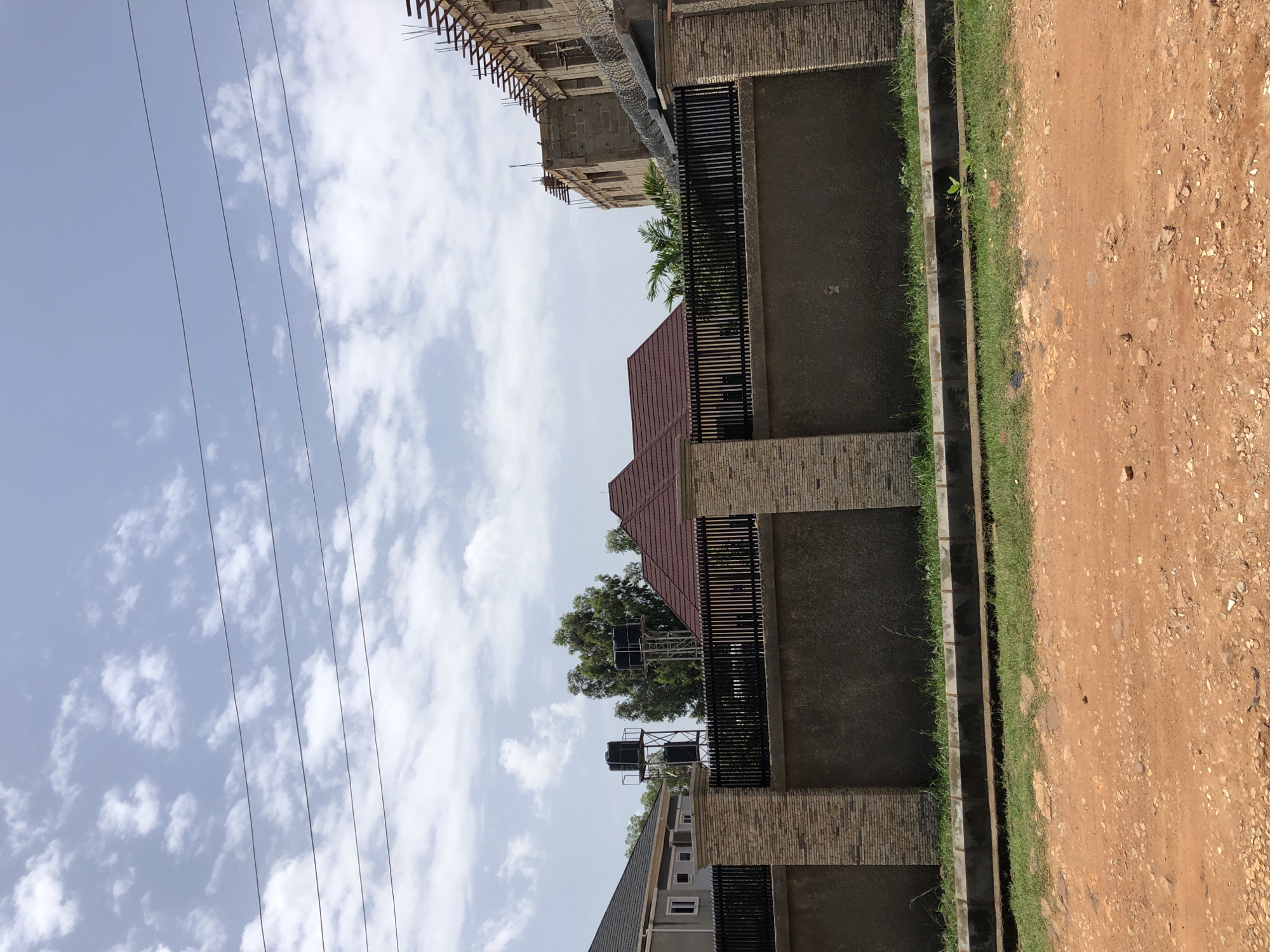 The image captures an outdoor scene in Bar Jirgi Summa, Nigeria. A light-colored, textured stone wall topped with a dark metal fence stretches across the foreground, separating a street from the properties beyond. Behind the fence, a dark red, gabled roof of a house is visible, partially obscured by trees. To the left of this house, a lighter-colored building with a dark roof and white-framed windows is present. Further back, two tall metal structures support black water tanks. To the right, a building under construction with exposed concrete blocks and scaffolding is partially visible. The sky above is blue with scattered white clouds, and several parallel wires run horizontally across the upper portion of the frame. The ground in the immediate foreground appears to be reddish-brown dirt and gravel.