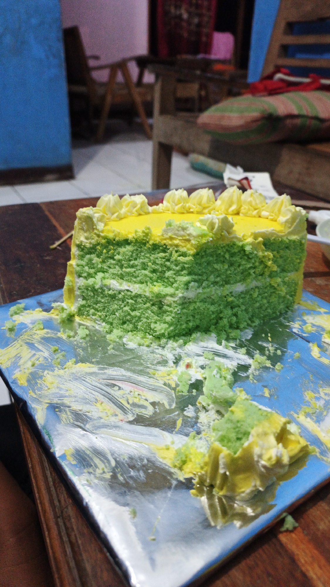 A slice of green and yellow layered cake with yellow frosting swirls on top sits on a blue foil tray on a wooden table. The cake has been partially eaten, with crumbs and frosting scattered on the tray and the table. In the background, a wooden chair with a striped cushion is visible. The environment appears to be indoors, possibly a home kitchen in Gunungreja, Indonesia, given the context.