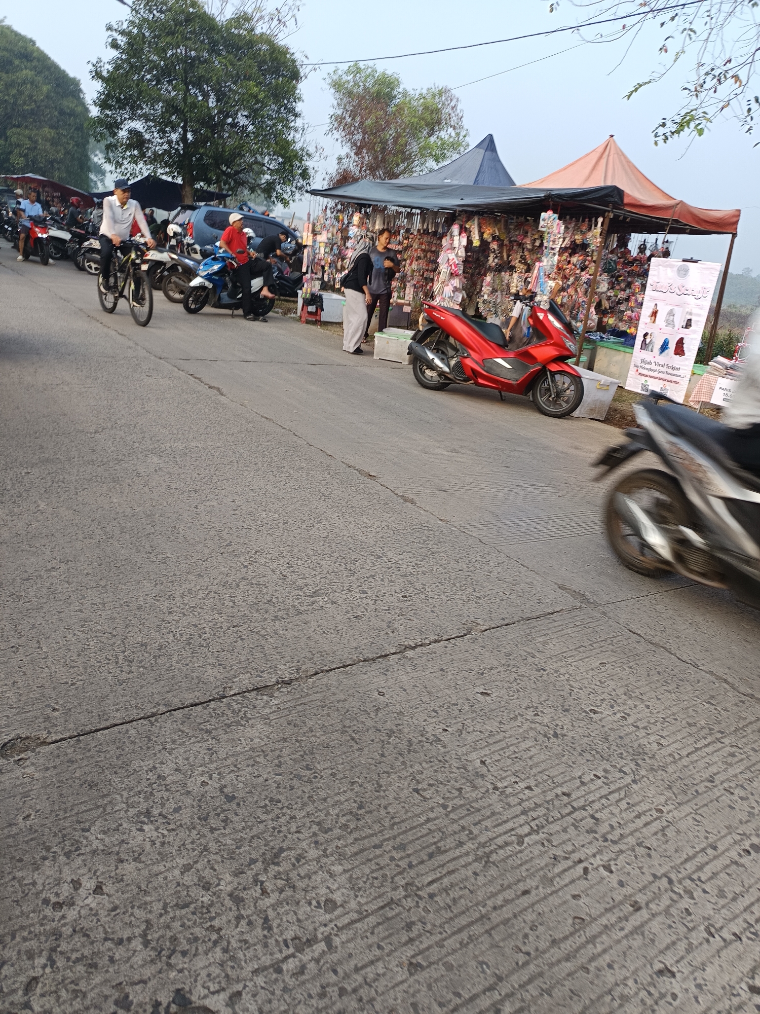 The image depicts a street scene in Sodong, Indonesia, characterized by a bustling roadside market and vehicular activity. In the foreground, a gray asphalt road shows distinct parallel grooving. To the left, a cyclist in a white shirt and blue cap rides a bicycle, his legs in motion. Behind him, several motorcycles are parked, with some riders visible. A blue SUV is parked further back. The central and right portions of the image are dominated by a roadside market with two tents, one orange and one dark blue, providing shade. The stalls are filled with an assortment of merchandise, including numerous keychains and other small items hanging from display racks. A bright red scooter is parked in front of the stalls. A sign in the foreground advertises "Hijab Viral Terkini," suggesting clothing or accessories for sale. Several individuals are browsing or working at the market. A person in a white shirt and black pants stands near the red scooter, and another individual in a black shirt and white pants is seen to their right. Further down the road, another motorcycle, blurred due to motion, is visible. The environment appears to be outdoors, with trees lining the background, suggesting a semi-rural or suburban setting on the outskirts of Sodong. The sky is lightly overcast.