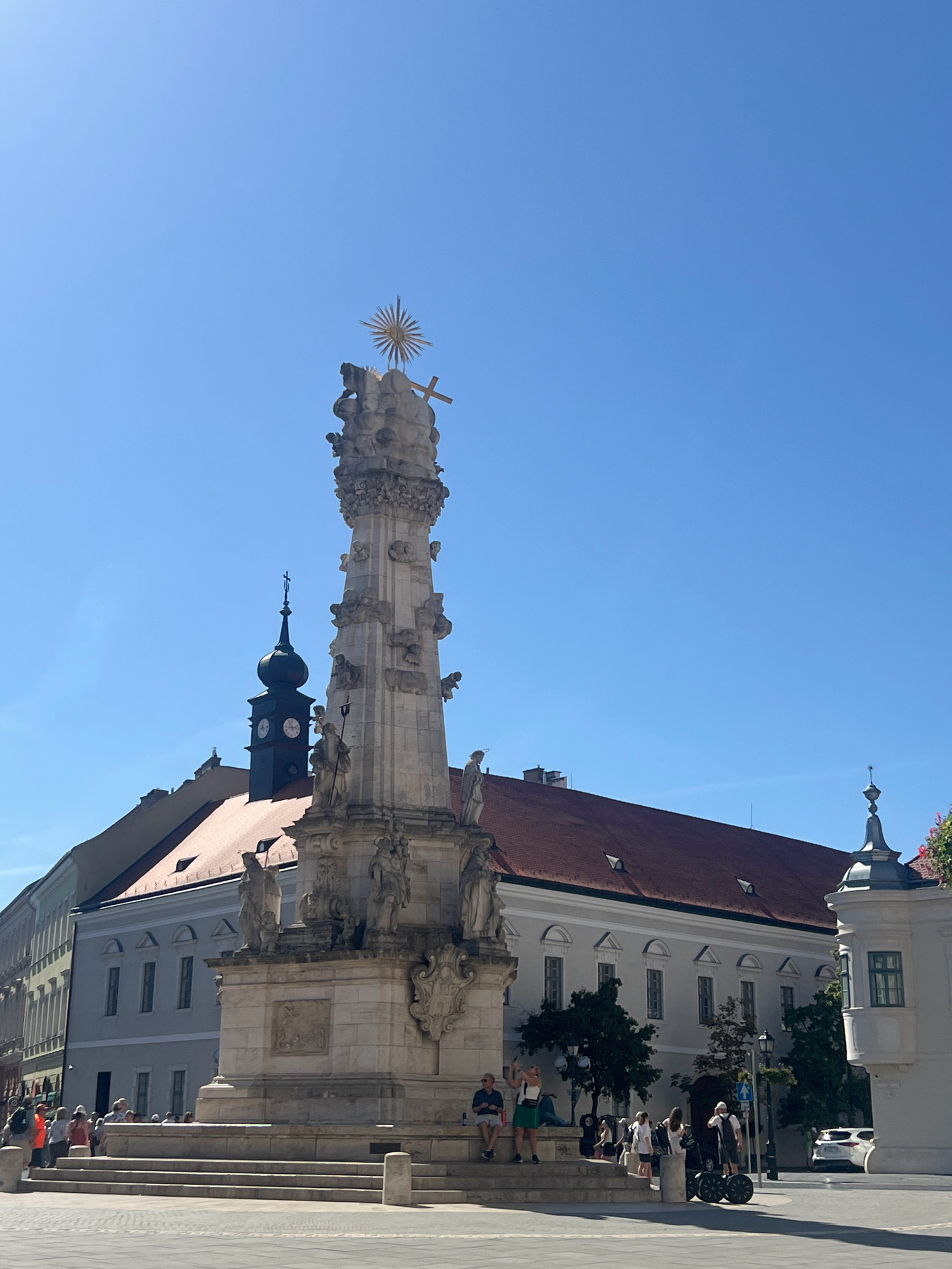 The image depicts a plague column in a European city square on a sunny day. The column, made of white stone, is elaborately decorated with statues of saints and intricate carvings. At the top, it is crowned with a gilded sunburst and a cross. In the background, several buildings with red tile roofs and light-colored facades line the square. A black clock tower with a steeple is visible behind the column. The ground is paved with light-colored cobblestones. A number of people are present in the square, some sitting on the steps of the column, while others are standing or walking. One person is riding a Segway. The sky is clear and bright blue. The scene appears to be a public gathering space, likely a tourist attraction in a city with historical architecture. Based on the architectural style, the plague column, and the overall ambiance, the location is identified as Budapest, Hungary.