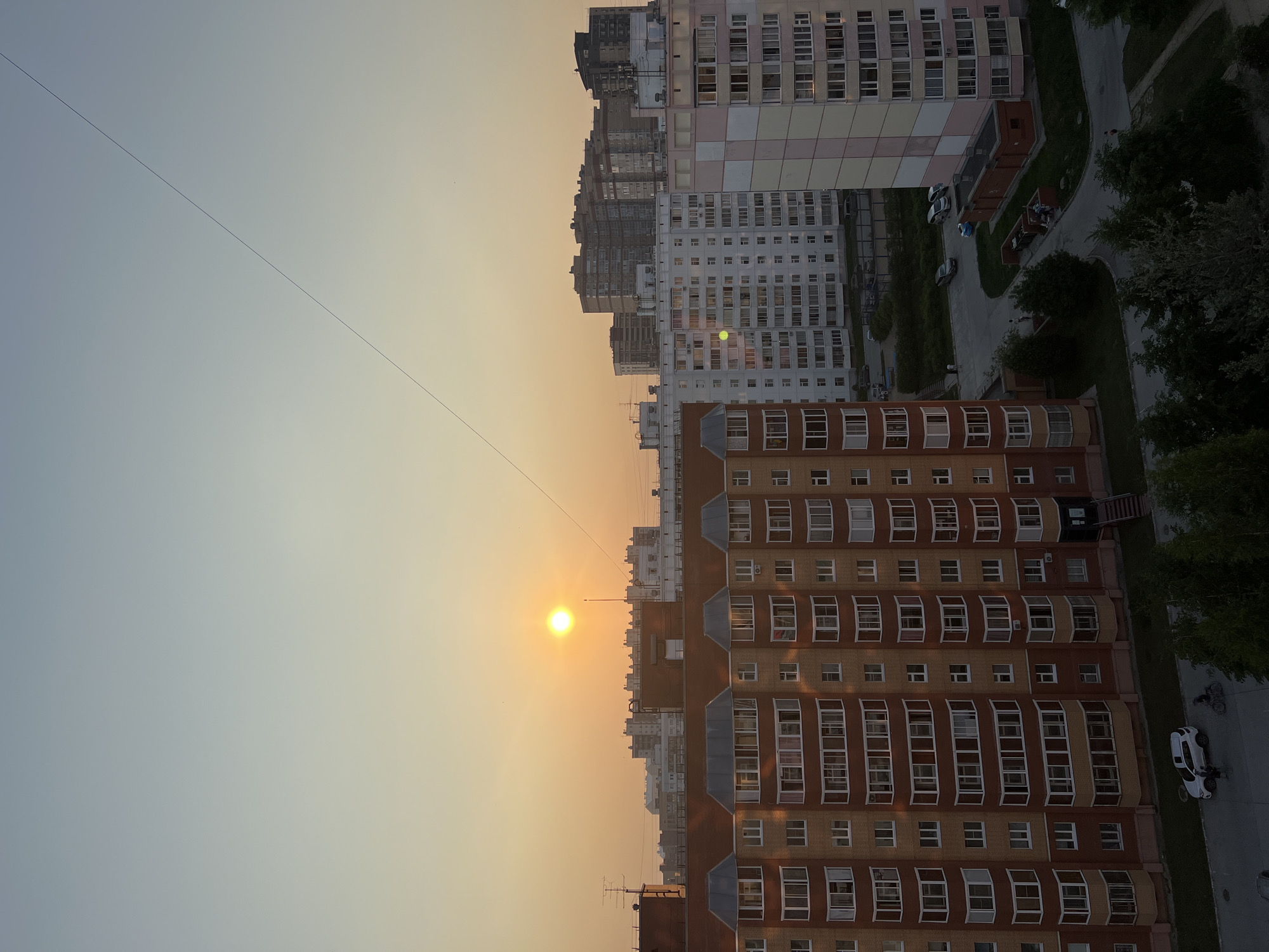 This image, taken in Novosibirsk, Russia, captures a cityscape at sunset. The scene is dominated by tall apartment buildings, characteristic of urban Russian architecture. A thin wire stretches diagonally across the sky, a common sight in many urban environments. The sun, a bright, warm orb, is low on the horizon, casting a soft, golden light that bathes the buildings and the sky in a hazy glow. This suggests it is either sunrise or sunset, with the warm tones leaning towards sunset.
Several cars are parked on a road or courtyard adjacent to one of the buildings, and a few people are visible, suggesting a typical evening in a residential area. The buildings themselves are multi-story structures with numerous windows, many of which appear to be enclosed balconies or conservatories, a common architectural feature in Russia. The buildings exhibit a range of colors and architectural styles, with some featuring a patchwork of pastel colors and others a more uniform brick or concrete facade.
The overall atmosphere is one of urban tranquility, with the setting sun creating a peaceful and somewhat nostalgic mood. The sky transitions from a clear, pale blue on the left to a warm, hazy orange on the right, where it meets the buildings. The presence of greenery in the form of trees peeking from behind the buildings adds a touch of nature to the urban landscape. No discernible text is visible in the image.