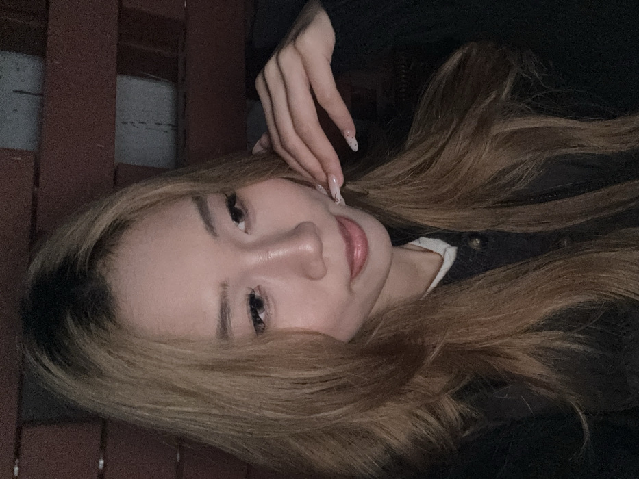 A close-up portrait captures a female individual in an indoor setting within Yuzhong District, China. The subject, appearing to be a young adult, possesses long, layered light brown hair with darker roots. She is clad in a dark outer garment over a light-colored collared shirt. Her left hand, adorned with long, light-colored nails featuring small dark dot patterns, is positioned with fingertips gently touching her left cheek. Her gaze is directed slightly downwards and to the right of the frame, accompanied by a subtle smile. The background is dimly lit and consists of horizontal reddish-brown panels or slats with lighter vertical elements or gaps.