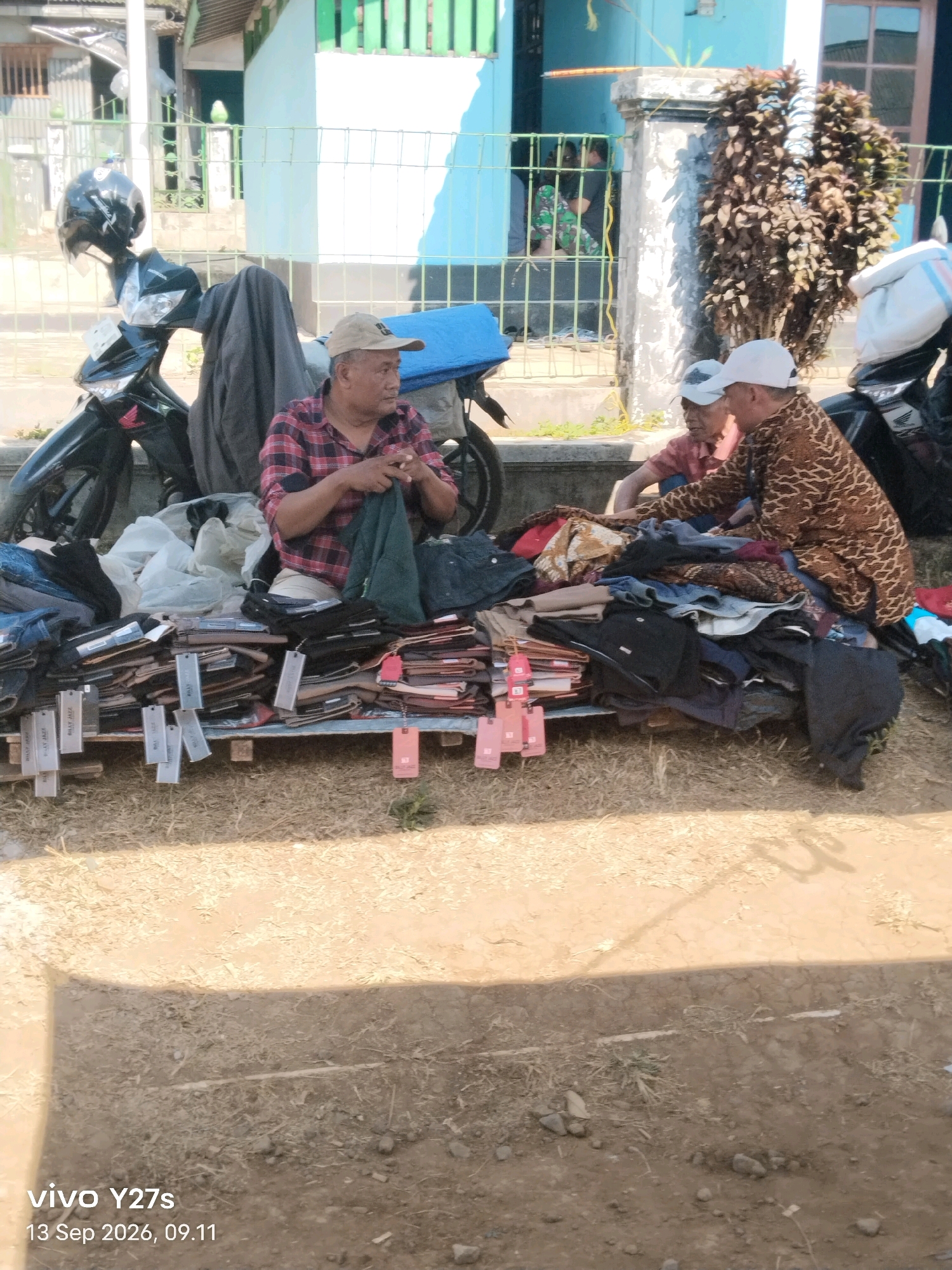 In Wonosido, Indonesia, two men are seen outdoors, appearing to be engaged in selling clothing. One man, wearing a red and black plaid shirt and a light-colored cap, is seated and holding a green garment, possibly for display or inspection. He is positioned in front of a display of folded clothing. Beside him, another man, wearing a white cap and a patterned brown shirt, is also seated and looking towards the clothing. The scene is set on a dirt ground, with a variety of folded clothes spread out on wooden pallets or a similar surface. In the background, a motorcycle is parked, with a dark cloth draped over its handlebars. The setting suggests an open-air market or roadside stall.