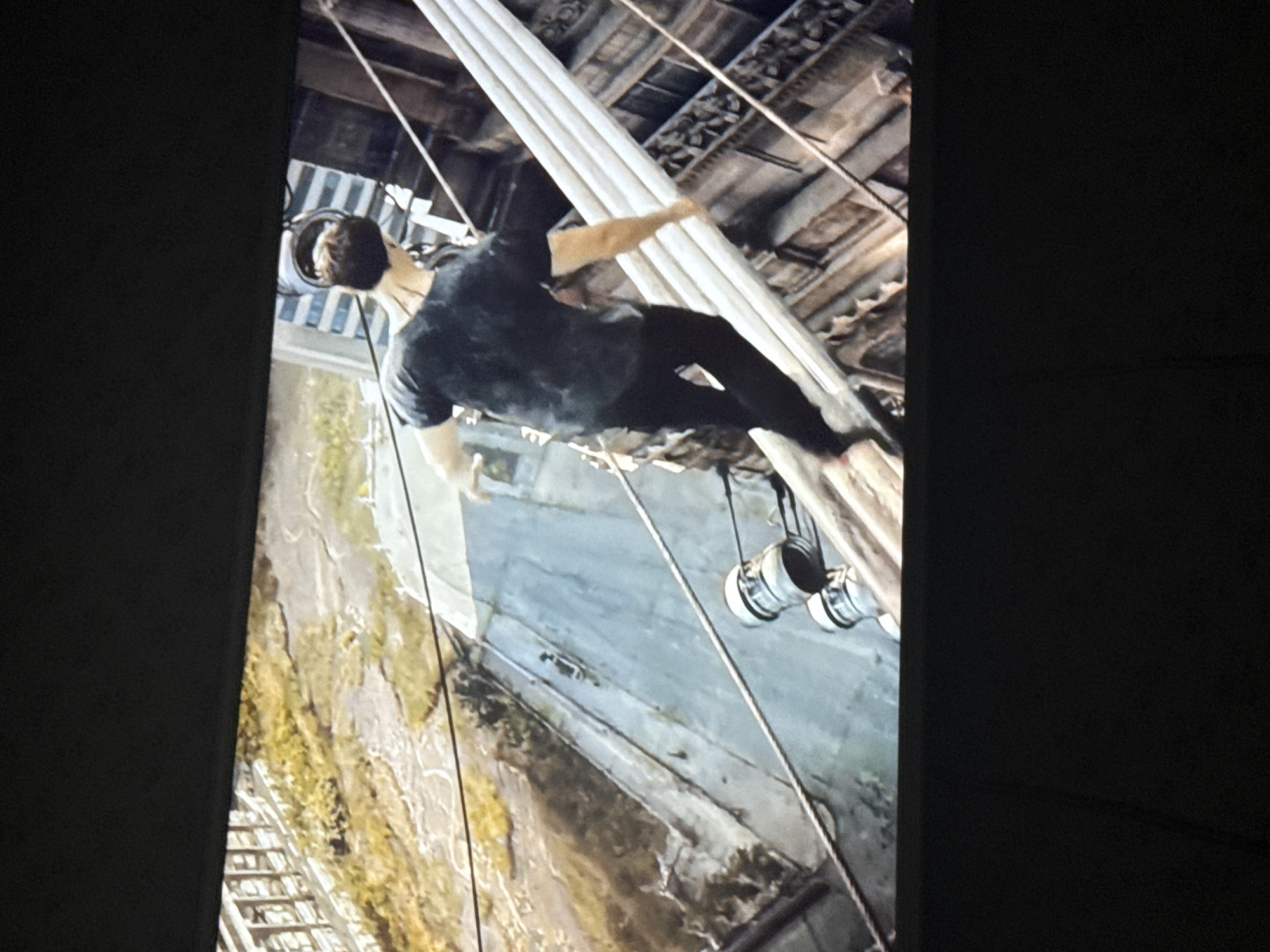 A man in a dark t-shirt and pants is scaling a tall, white structure resembling large pipes or beams. He is wearing headphones and appears to be suspended high above the ground. The backdrop includes elements of urban architecture, with visible building structures, and a view looking down at street-level details. Based on the location of Mytishchi, Russia, there is nothing in the photo that indicates it is located there.