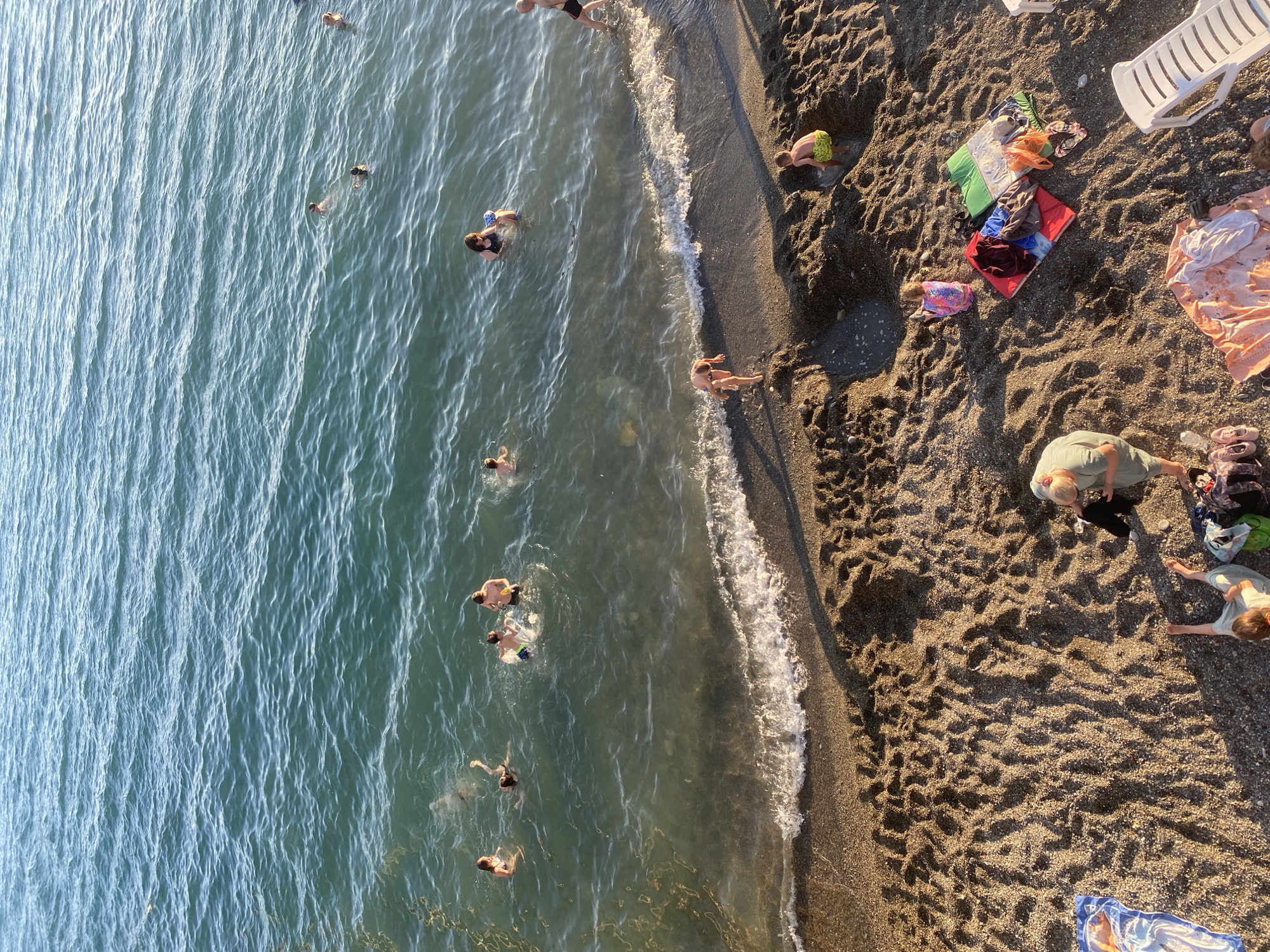 This is an aerial, high-angle shot of a crowded beach in Sochi, Russia. The image captures a lively scene with people enjoying a sunny day at the water's edge. The water is a clear, greenish-blue, showing gentle ripples and the movement of people swimming. Several individuals are visible in the sea, some swimming, others wading.
On the pebbly shore, a collection of colorful beach towels and blankets are spread out, indicating people relaxing or preparing to swim. There are also a few beach chairs, one white plastic chair prominently placed. Scattered items include bags and personal belongings. The sunlight casts long shadows across the beach, suggesting it's either late morning or late afternoon. The weather appears to be warm and clear, ideal for a day at the beach. The overall mood is one of leisure and enjoyment, with people engaged in typical beach activities. No visible text is present in the image.