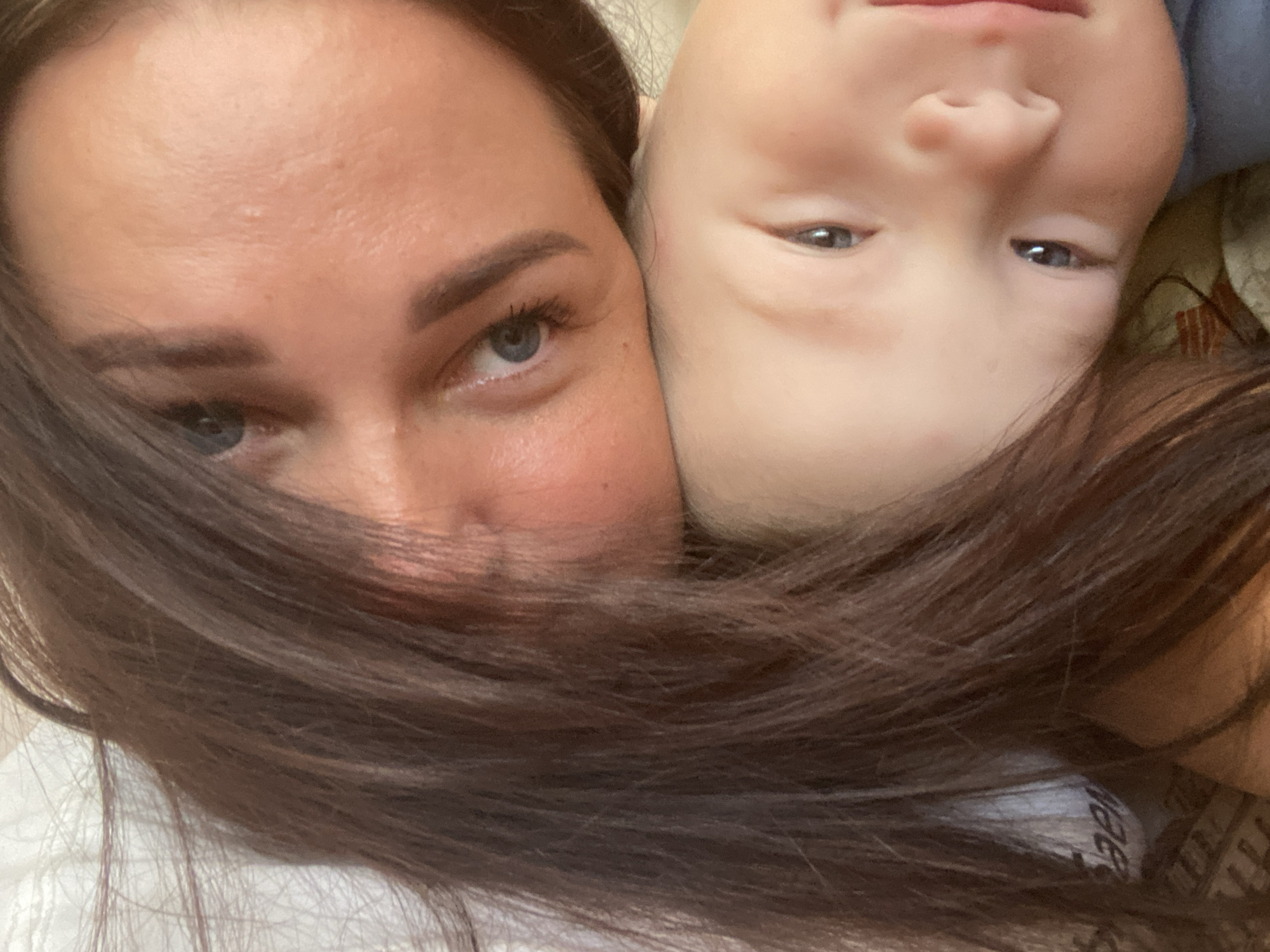 The image is a close-up, overhead shot of two individuals, likely an adult and a child, resting together. The adult's head is positioned at the top of the frame, with their dark hair falling over the child's head and upper body. The adult's face is partially visible, showing one eye with blue irises and dark eyebrows, and the upper portion of their forehead. The child's face occupies the lower part of the frame, tilted upwards. Their skin appears fair, and their eyes are dark. The lighting suggests an indoor setting, possibly a bedroom, with soft light illuminating their faces. No specific landmarks or urban features are visible to indicate a location within the city of Sochi, Russia.