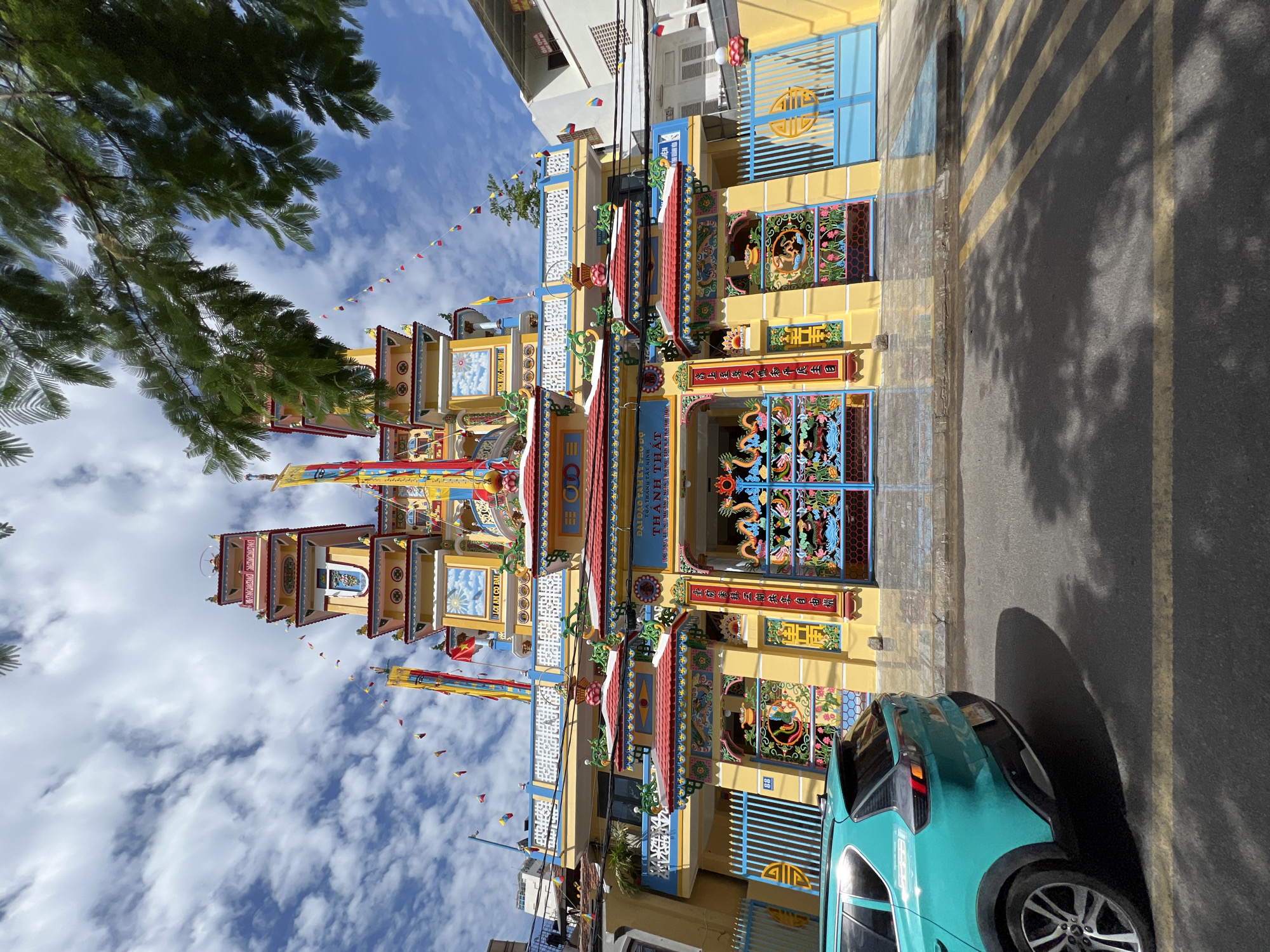 This is a ground-level, eye-level, slightly wide-angle outdoor shot of the ornate, vibrant facade of a Buddhist temple. The temple is predominantly yellow with red and blue accents and features multiple tiered roofs, decorative elements like dragons and lotus flowers, and flags. In the foreground, the rear left side of a turquoise SUV is visible, parked on an asphalt street. The street has yellow lane markings. The sky is blue with scattered white clouds, and tree branches with green leaves overhang the top right of the frame, casting shadows on the temple and street. The architecture suggests it is located in Southeast Asia, likely Vietnam, given the style and possible inscriptions on the temple. The inscription on the temple appears to be in Vietnamese or Chinese characters. The overall atmosphere is bright and sunny. This specific temple is identified as the **Long Sơn Pagoda (Long Son Tu)** in **Nha Trang, Vietnam**.