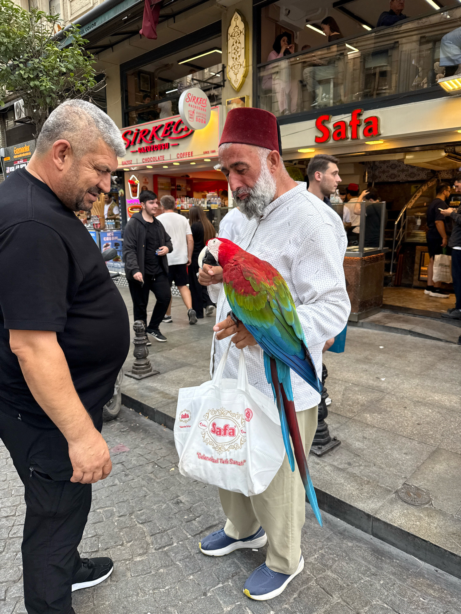 In an outdoor, urban setting, two men stand on a cobblestone street. The man on the left, wearing a black t-shirt and black pants, has his side to the camera, with his left arm bent at the elbow and his hand resting on his stomach. He has gray hair and a beard. The man on the right, facing the camera, wears a white patterned shirt, a red fez, and tan pants. He holds a colorful parrot with red, green, and blue plumage on his left hand. He is also holding a white shopping bag with red text that reads "Safa". Behind them, a store with a red neon sign that says "Sirkecci" is visible, and further back, another store sign reads "Safa". Other pedestrians are visible in the background, some walking and some looking into shop windows. The overall scene appears to be a bustling street in a city.