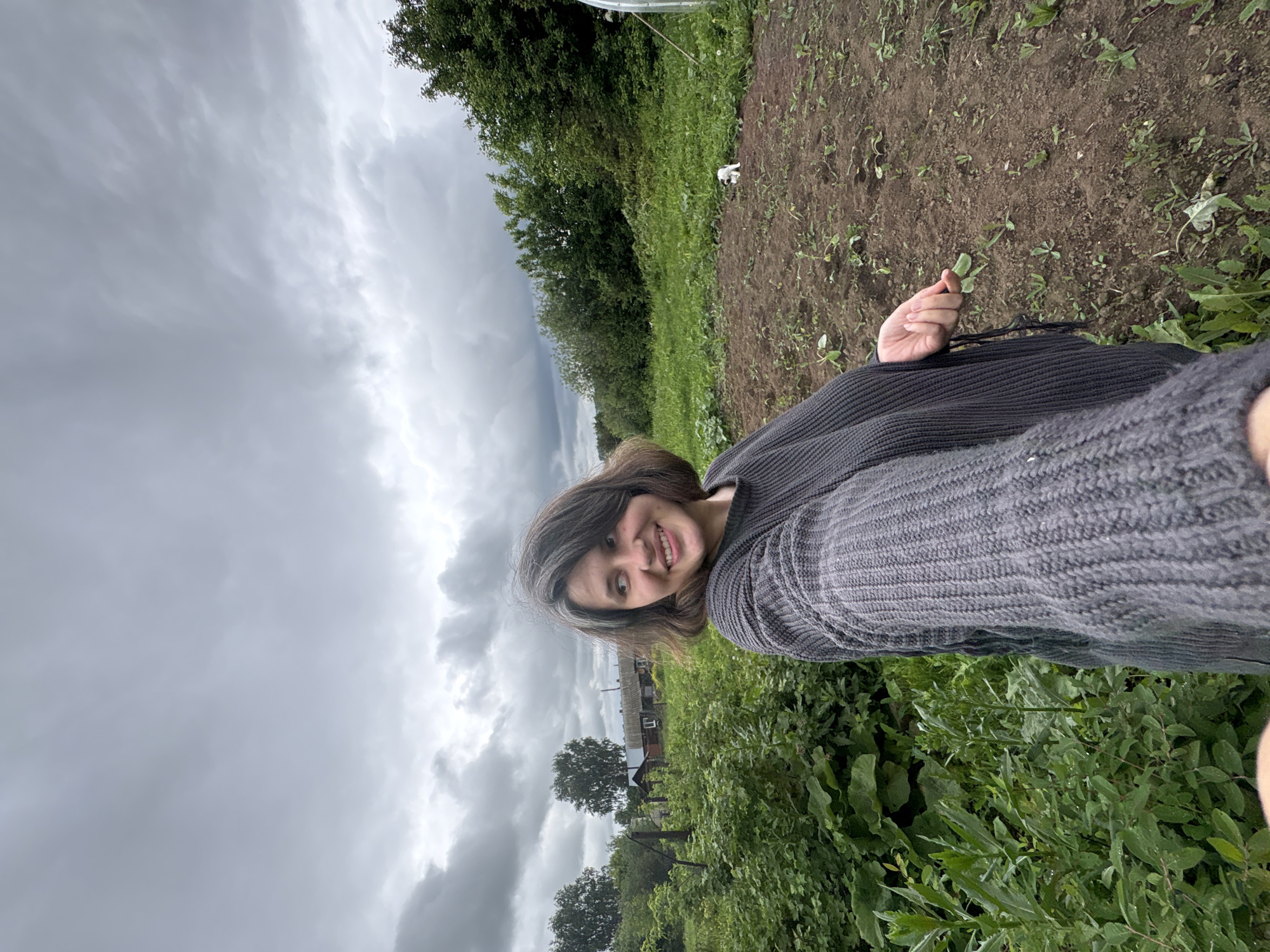 An adult female is depicted in a rural outdoor setting under a heavily overcast sky in the city of Фоки, Russia. She is positioned in the foreground, facing slightly right from the viewer's perspective, with her head turned further to her left (viewer's right). She has shoulder-length, dark brown hair with visible grey strands and a light complexion, displaying a broad smile with teeth visible. She wears a chunky, dark grey knitted sweater with long sleeves. Her right arm is extended towards the camera, suggesting a selfie perspective. Her left hand is visible, holding a small green leaf or plant sprout.
The immediate foreground on the left consists of dense green foliage, including various leafy plants. A wooden fence post is partially visible within this greenery. On the right, a patch of tilled, dark brown earth, likely a garden bed, contains sparse, small green plant sprouts. A small, white and black object is visible on the edge of this cultivated patch, near a transition to more grass and wild greenery.
In the midground, beyond the garden, a mix of green grass and uncultivated plants extends. The background features several wooden houses with dark roofs, partially obscured by trees. A dense treeline composed of various deciduous trees, including some with lighter bark, forms the horizon. The sky is entirely covered by thick, grey clouds, indicating overcast weather conditions.
