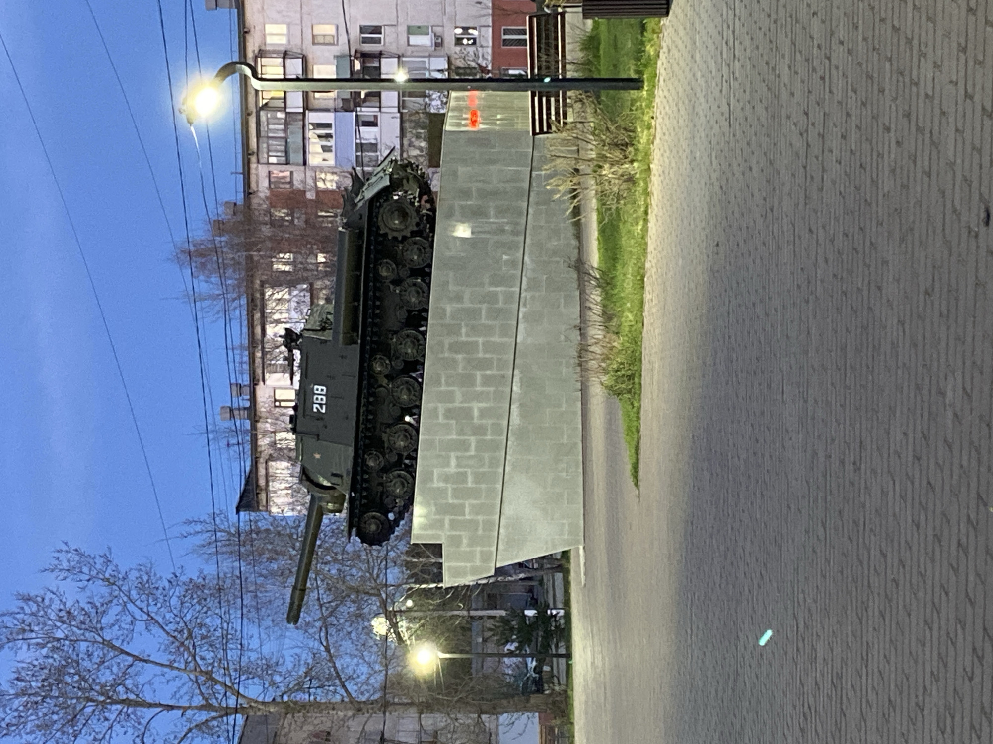The image depicts a military tank monument in Chaikovsky, Russia. The tank, painted in a dark green color, is mounted on a concrete pedestal made of gray blocks. The number "288" is prominently displayed on the side of the tank. The monument is located on a paved walkway, adjacent to a grassy area. A streetlight illuminates the scene. In the background, a multi-story apartment building is visible, along with bare trees silhouetted against the early evening sky. The perspective of the image is rotated.