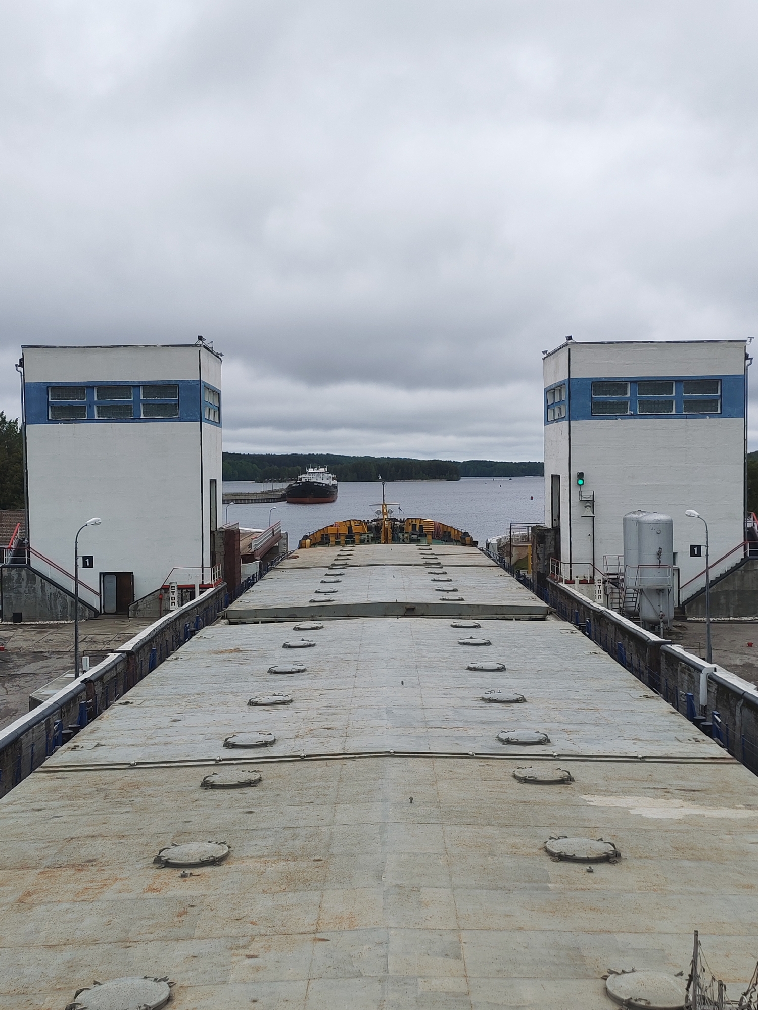 The image depicts a shipping canal lock in Белоусово, Russia. Two white buildings with blue accents flank either side of the canal. A large cargo ship is seen further down the canal, approaching the lock. The foreground is dominated by a gray, flat surface with circular hatches, presumably the top of the lock mechanism. The sky is overcast. There are no people visible in the image.