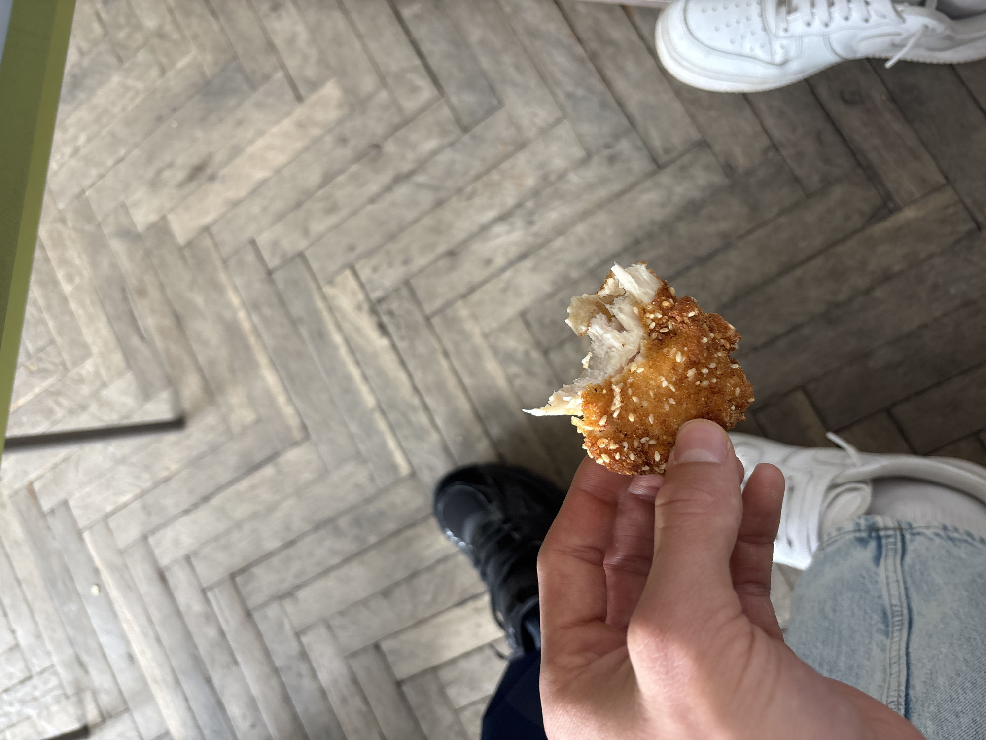 The image depicts a person holding a partially eaten sesame-crusted fried food item, possibly chicken, in their right hand. The person is wearing light blue jeans and white sneakers. The background shows a herringbone patterned floor, and two different shoes: one black and one white. Location is unknown.