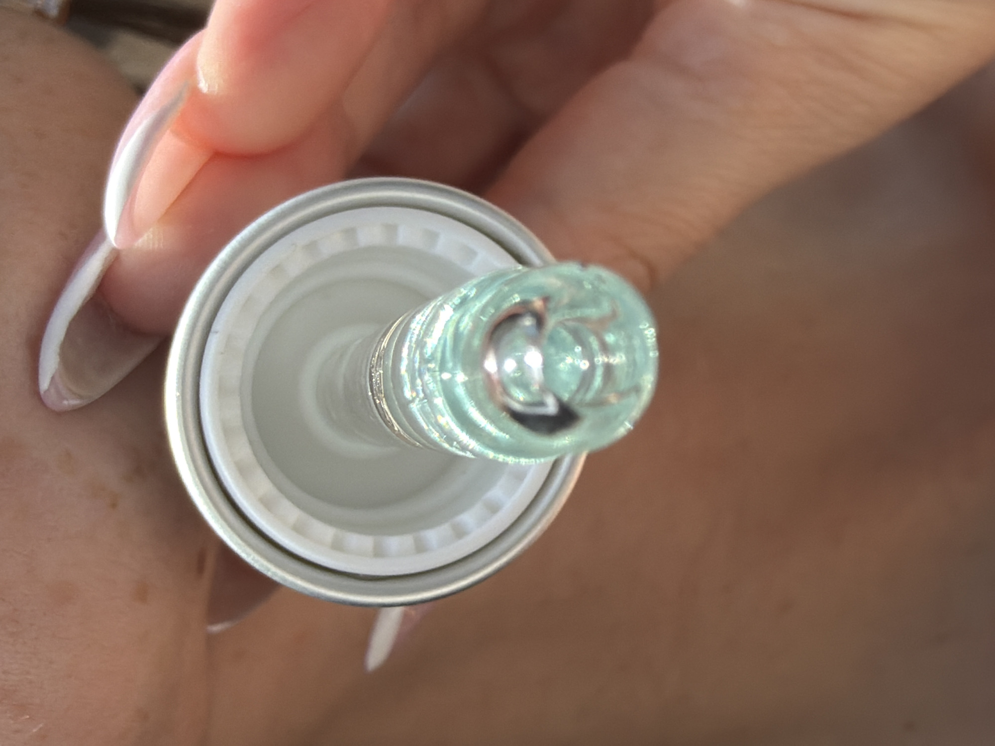 A person with long, white-tipped nails is holding a bottle of clear liquid with a silvery cap. The liquid appears to be a light blue or green tint.