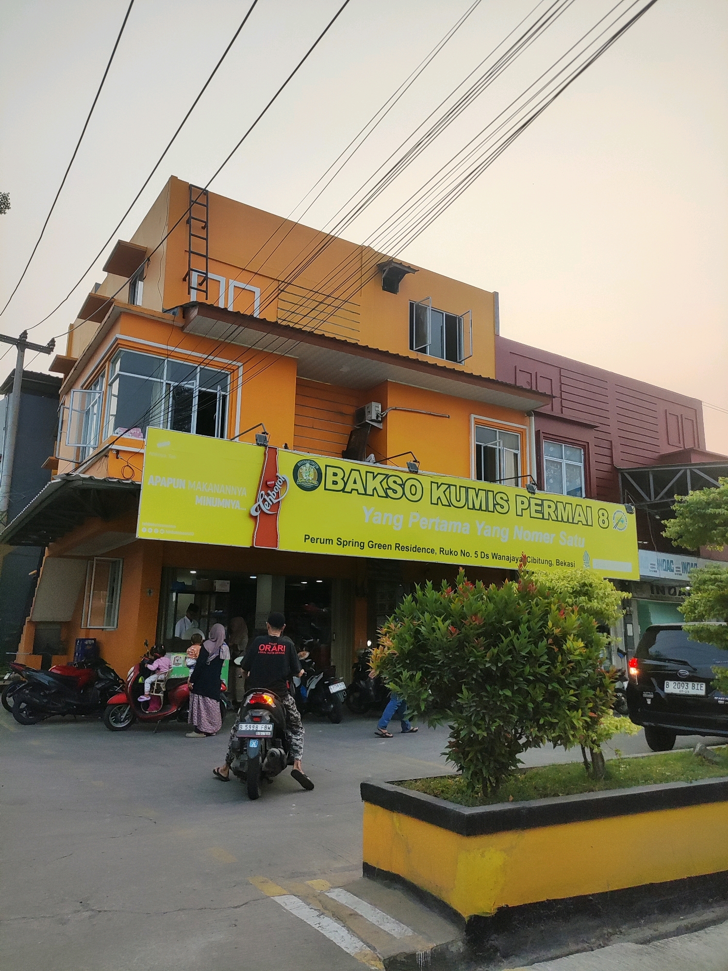 The image shows a two-story building with an orange facade, located in Wanajaya, Indonesia. A prominent yellow banner displays "BAKSO KUMIS PERMAI 8" and advertising for Teh Botol, along with an address in Cibitung, Bekasi. Multiple motorcycles are parked in front of the building, and several people are visible, some of whom appear to be customers. A person is riding a motorcycle in the foreground. The sky is overcast with electrical wires visible against it. The scene depicts a street-level view of a commercial establishment, likely a restaurant, with activity on the sidewalk and street.