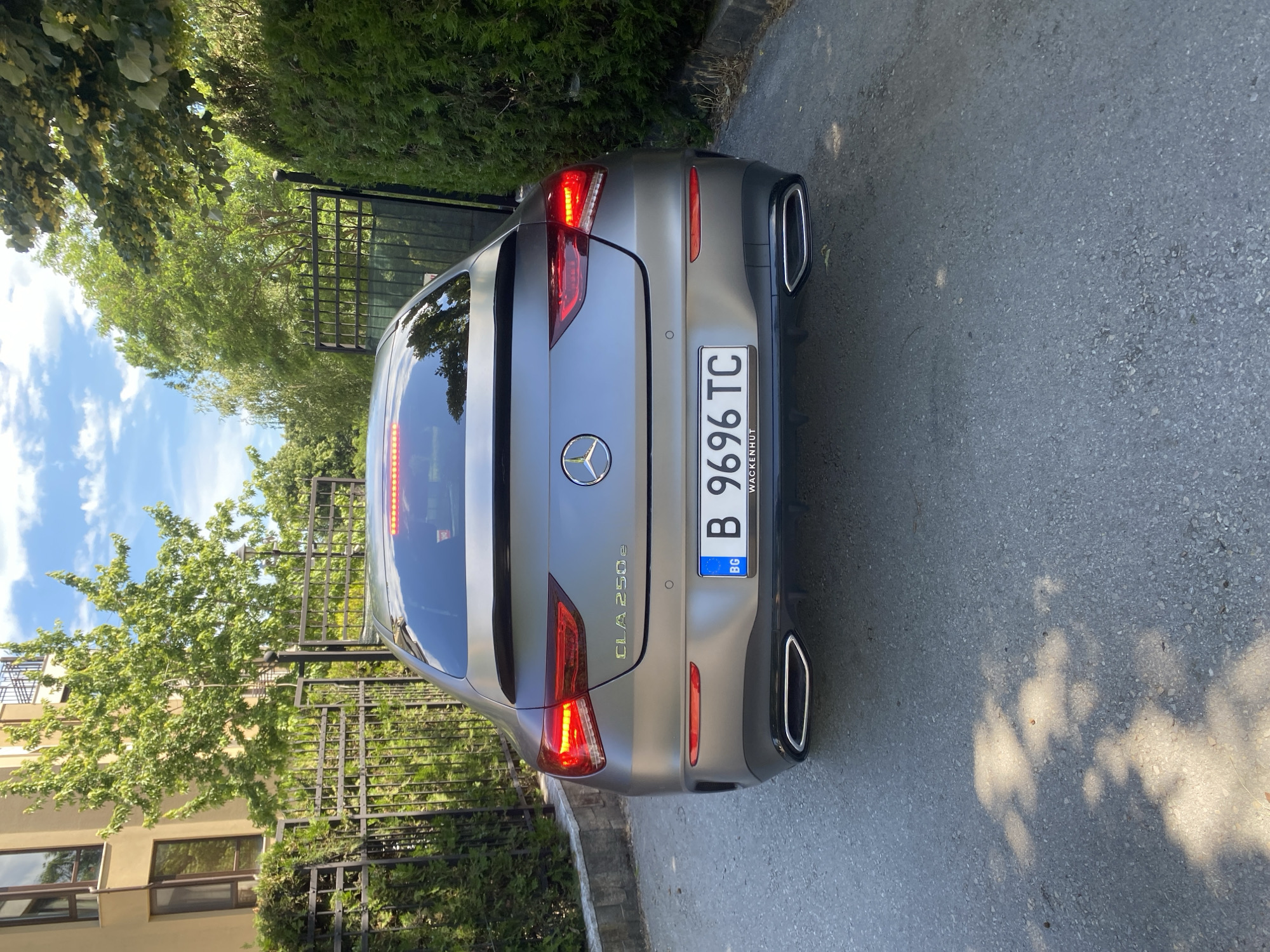The media file shows the rear view of a gray Mercedes-Benz CLA 250e parked on an asphalt road. The car is positioned at an angle, with the left side closer to the viewer. The background features a black metal fence, lush green trees, and a building with windows. The sky above is blue with scattered white clouds.
The car's taillights are illuminated, suggesting it might be evening or the lights are on for visibility. The license plate is visible and reads "B 9696 TC" with "BG" on a blue strip. There is also text below the license plate, partially obscured, but "WACKENHUT" is legible. The model name "CLA 250e" is also visible on the car's trunk.
The scene appears to be a residential area, possibly a driveway or a street next to a property. The sunlight suggests it is daytime, likely afternoon given the shadows cast on the road by the trees. The overall mood is calm and serene, with no people or animals present. The environment is well-maintained, with neatly trimmed foliage and a clean road surface.